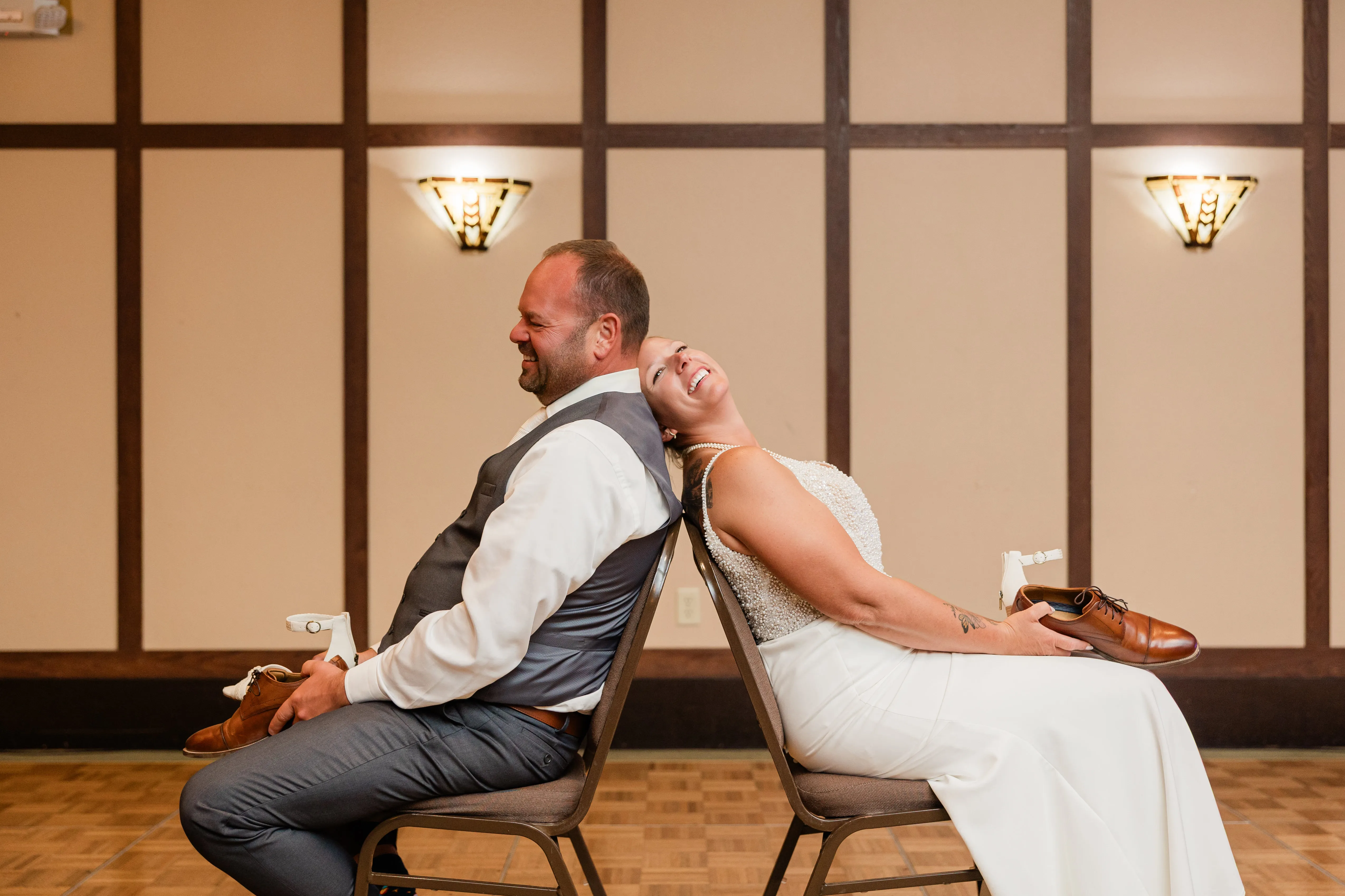 The bride and groom sit back-to-back on chairs during the shoe game at their reception, each holding a shoe and laughing as she leans her head against his shoulder — Tim Larsen Photography, Brainerd Lakes MN