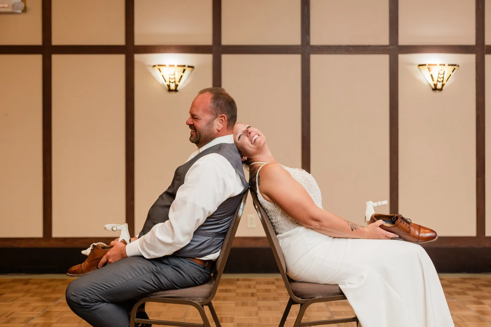 The bride and groom sit back-to-back on chairs during the shoe game at their reception, each holding a shoe and laughing as she leans her head against his shoulder — Tim Larsen Photography, Brainerd Lakes MN