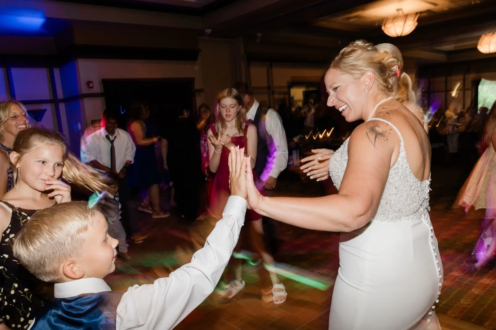 The bride laughs as she meets a young boy's raised hand in a high-five on a crowded, dimly lit dance floor surrounded by blurred dancing guests at Chase on the Lake — Tim Larsen Photography, Brainerd Lakes MN