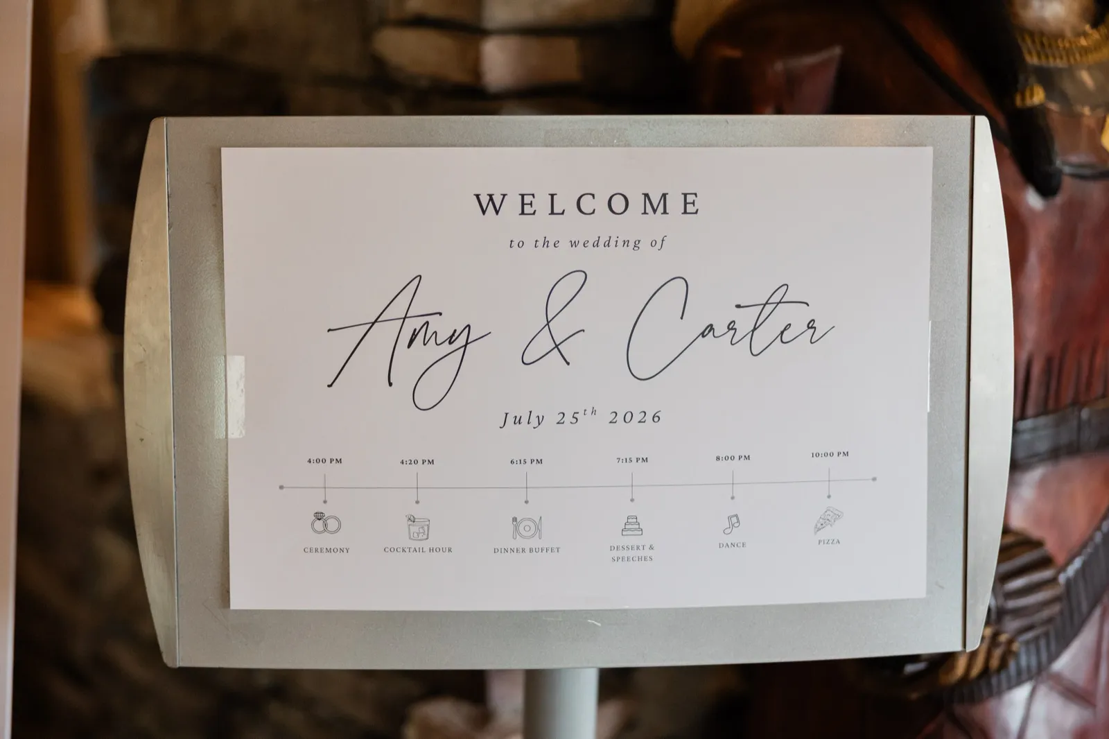 A white welcome sign on a metal easel lists Amy and Carter's July 25 wedding timeline against the stone wall inside Whitefish Lodge — Tim Larsen Photography, Brainerd Lakes MN