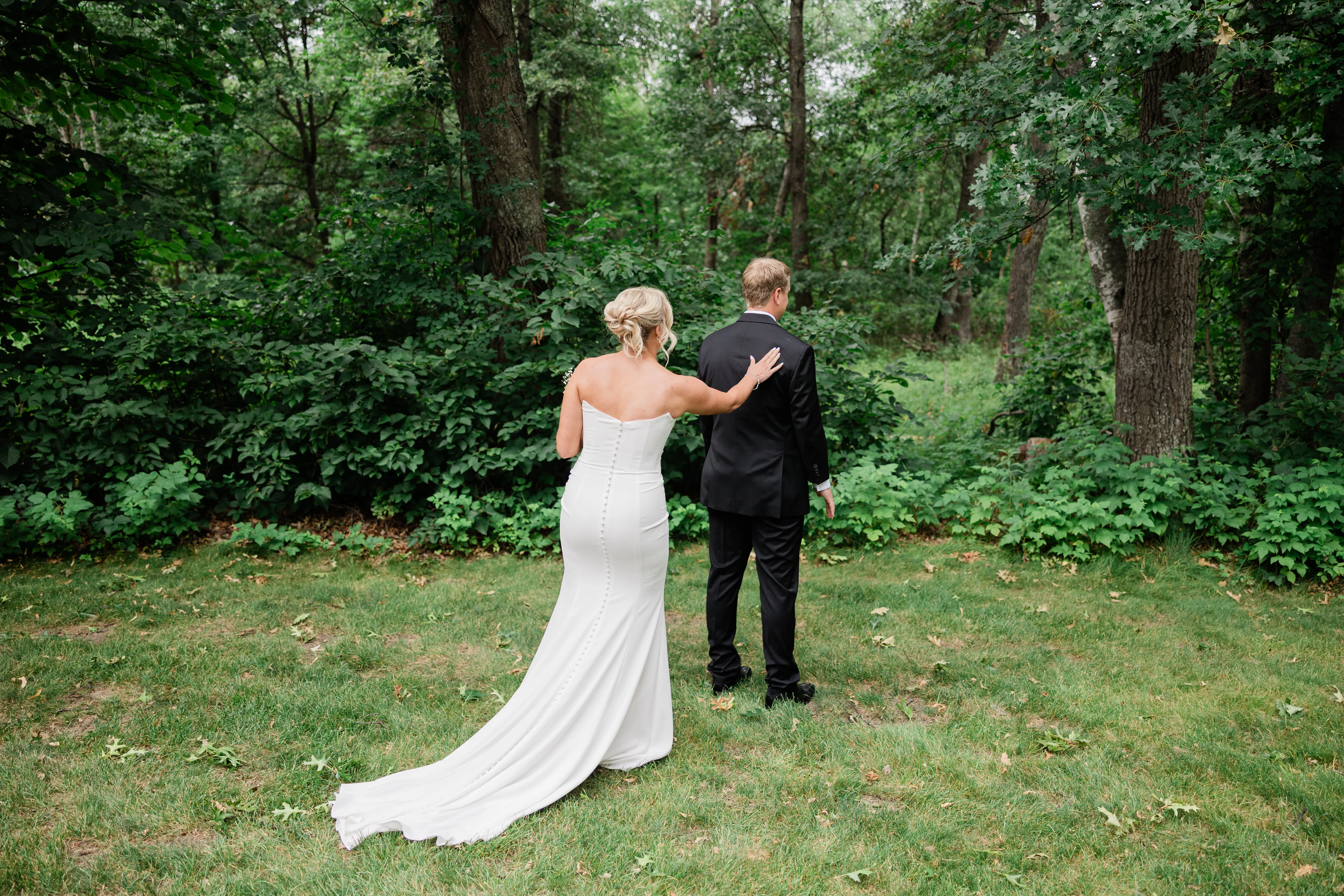 Amy reaches from behind to touch Carter's shoulder for their first look in a green wooded clearing, her train trailing across the grass — Tim Larsen Photography, Brainerd Lakes MN