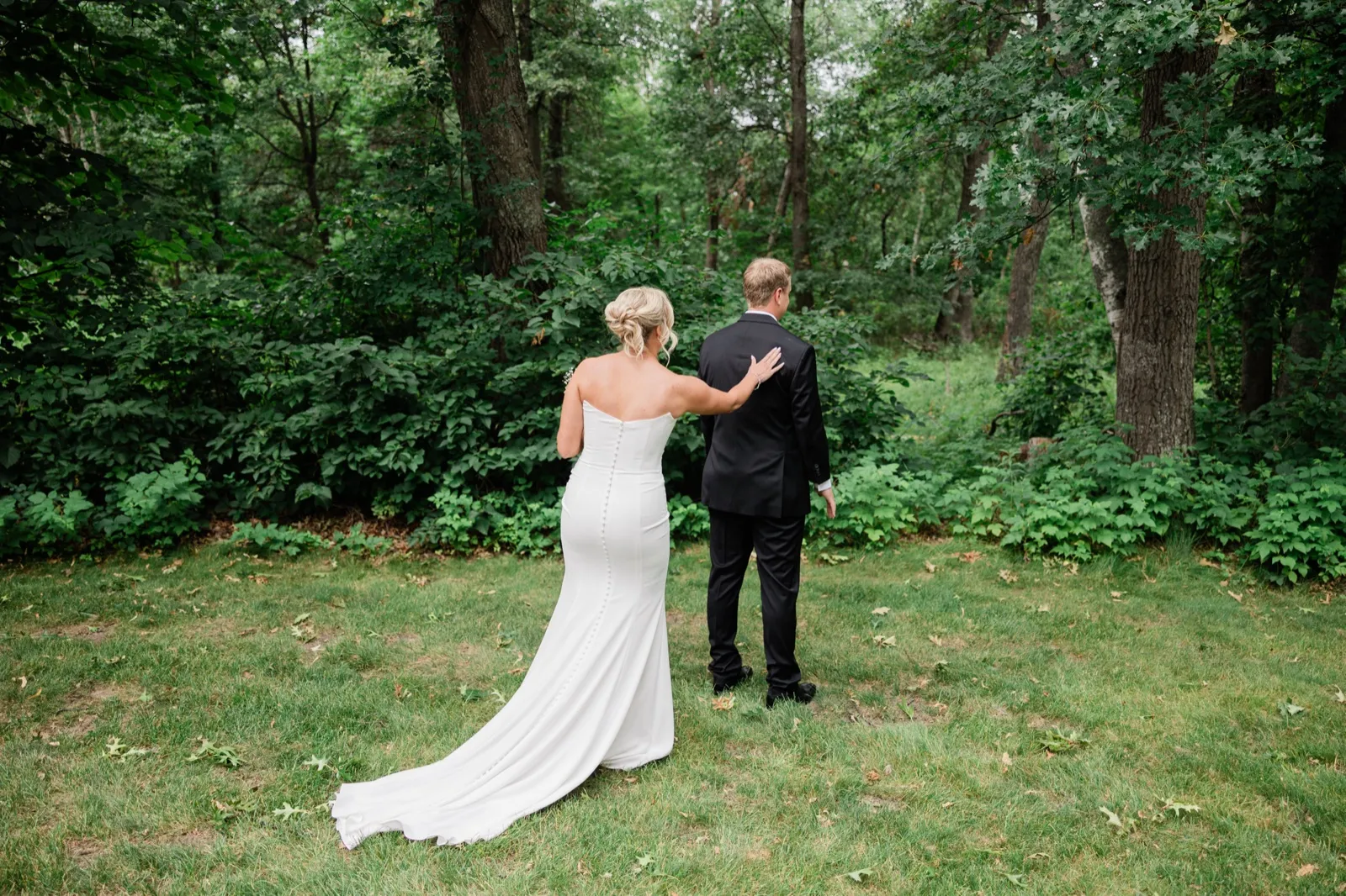 Amy reaches from behind to touch Carter's shoulder for their first look in a green wooded clearing, her train trailing across the grass — Tim Larsen Photography, Brainerd Lakes MN