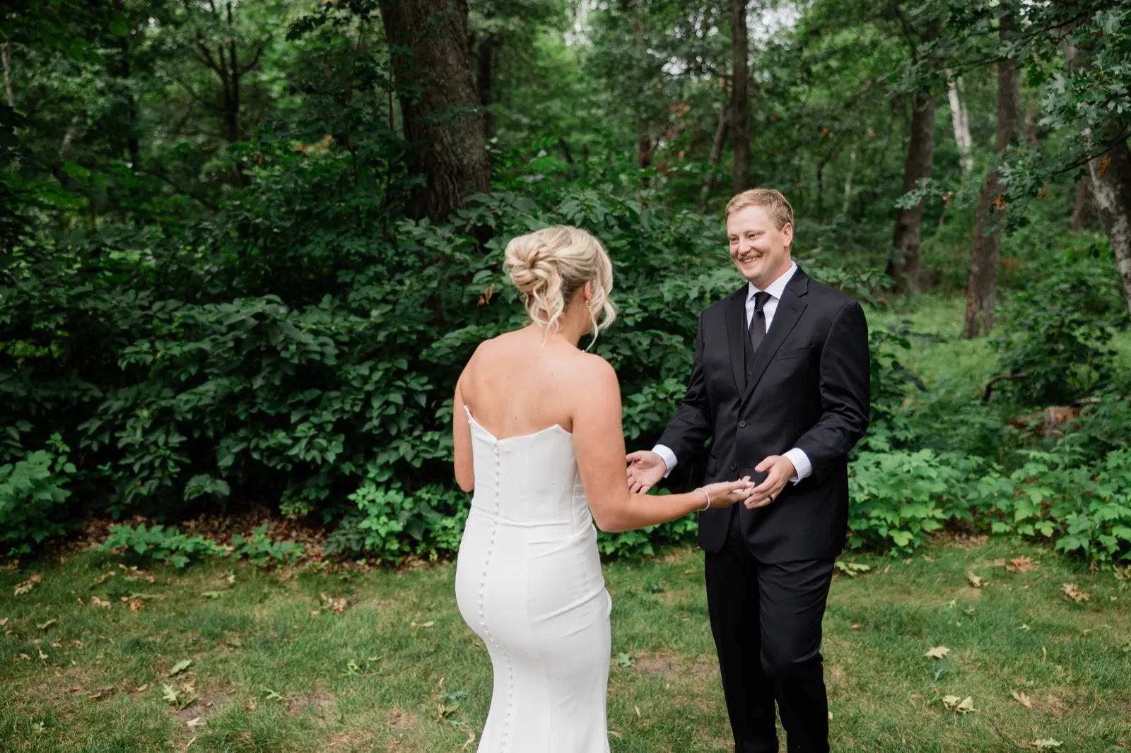 Carter turns and grins at Amy during their first look in the woods behind Whitefish Lodge, reaching for her hands — Tim Larsen Photography, Brainerd Lakes MN