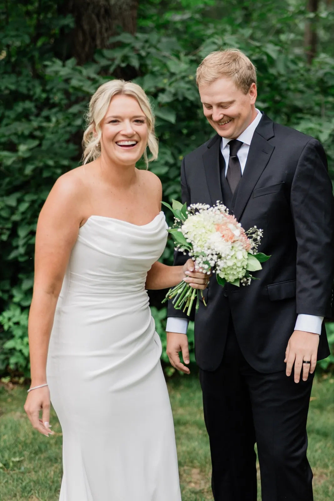 Amy laughs toward the camera while Carter looks down at her bouquet, the two framed by dense green summer foliage — Tim Larsen Photography, Brainerd Lakes MN
