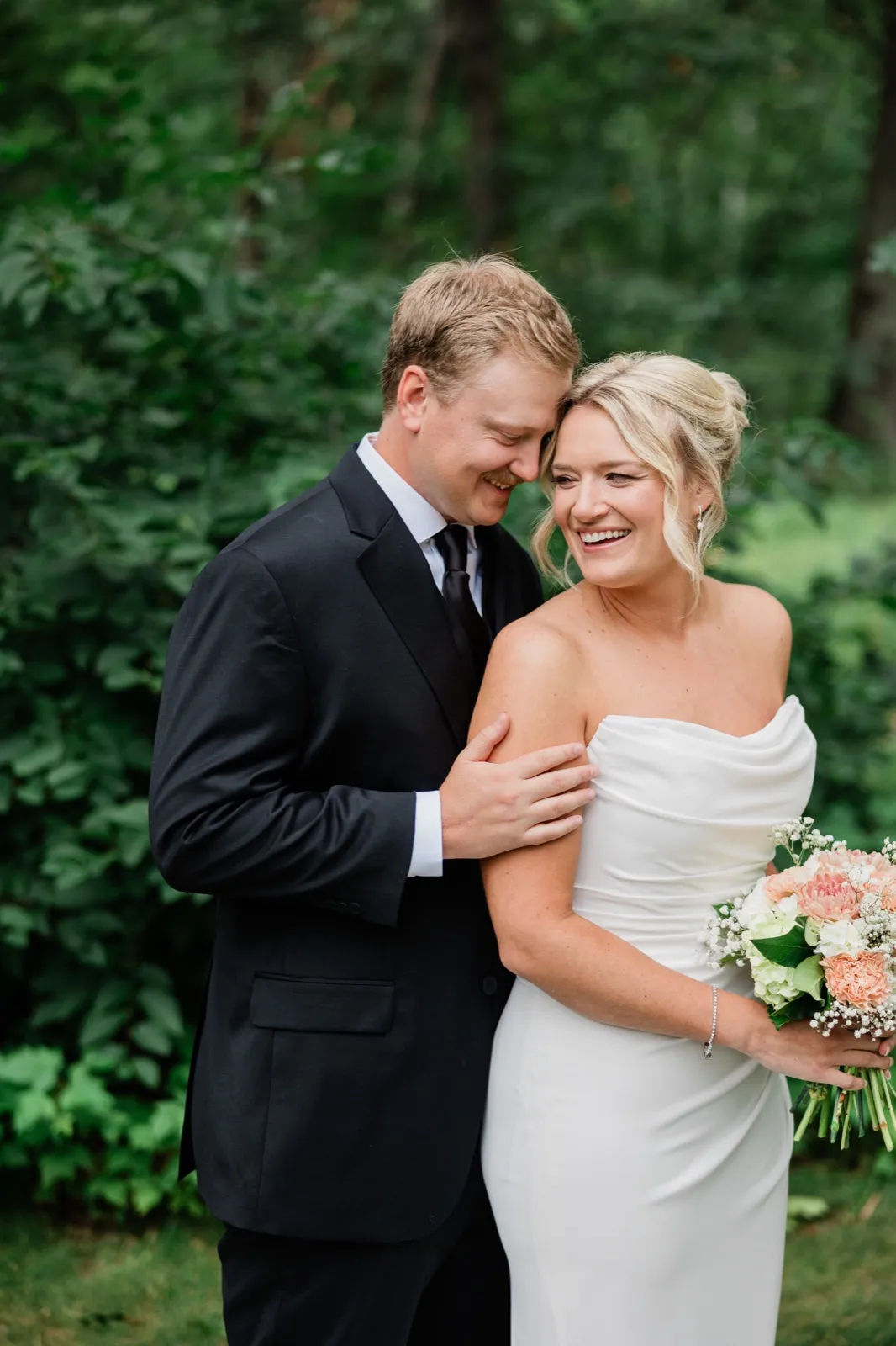 Carter leans his forehead toward Amy's temple as she laughs, both against dense green woods at Whitefish Lodge — Tim Larsen Photography, Brainerd Lakes MN