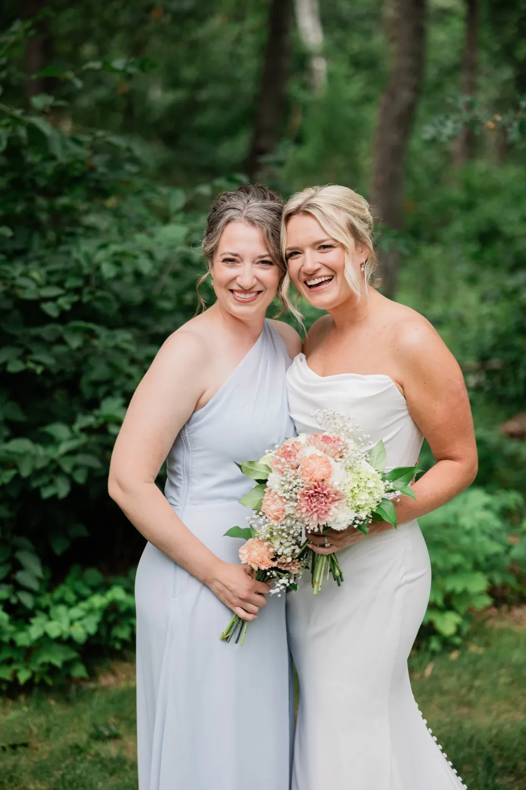 Amy leans cheek to cheek with an attendant in a pale blue dress, both laughing and holding peach and white bouquets — Tim Larsen Photography, Brainerd Lakes MN