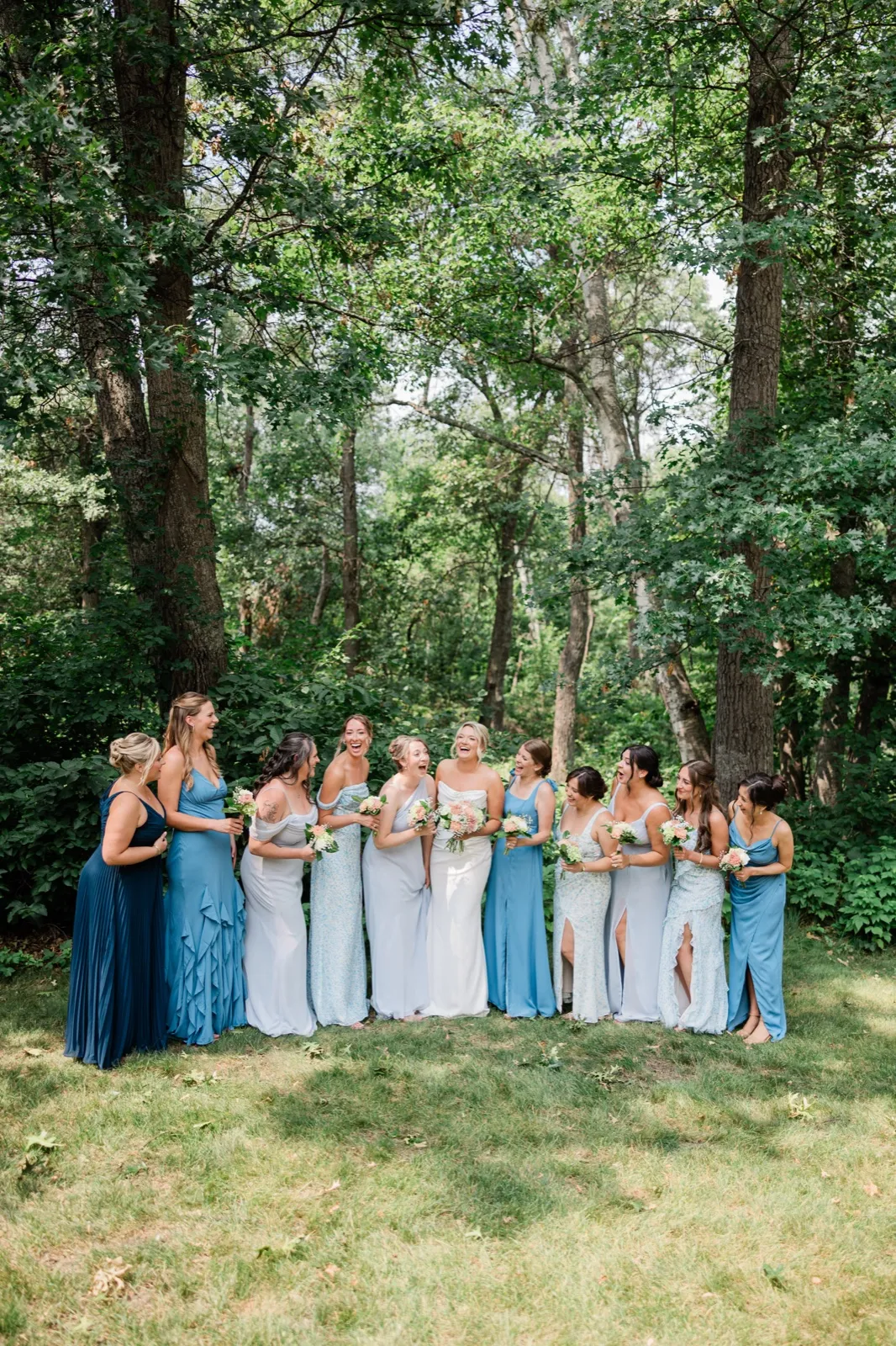Amy stands in white at the center of eleven women in every shade of blue, all laughing, beneath tall oaks at Whitefish Lodge — Tim Larsen Photography, Brainerd Lakes MN