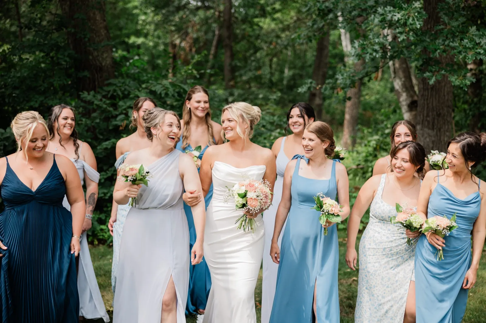 Amy walks arm in arm with her attendants in shades of blue across the grass in front of dense summer woods — Tim Larsen Photography, Brainerd Lakes MN