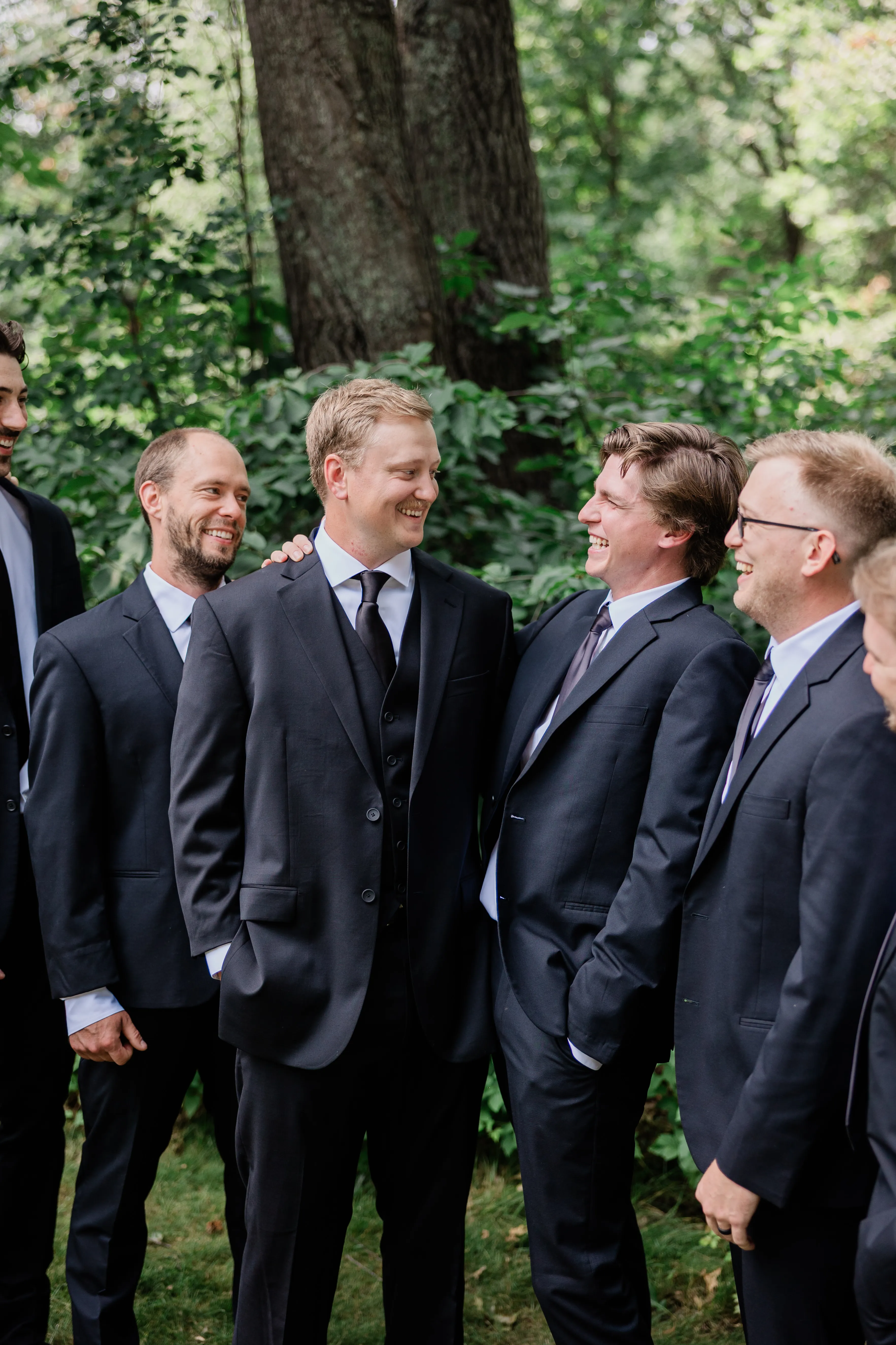 Carter laughs among five men in black suits who turn toward him, green summer woods behind them — Tim Larsen Photography, Brainerd Lakes MN