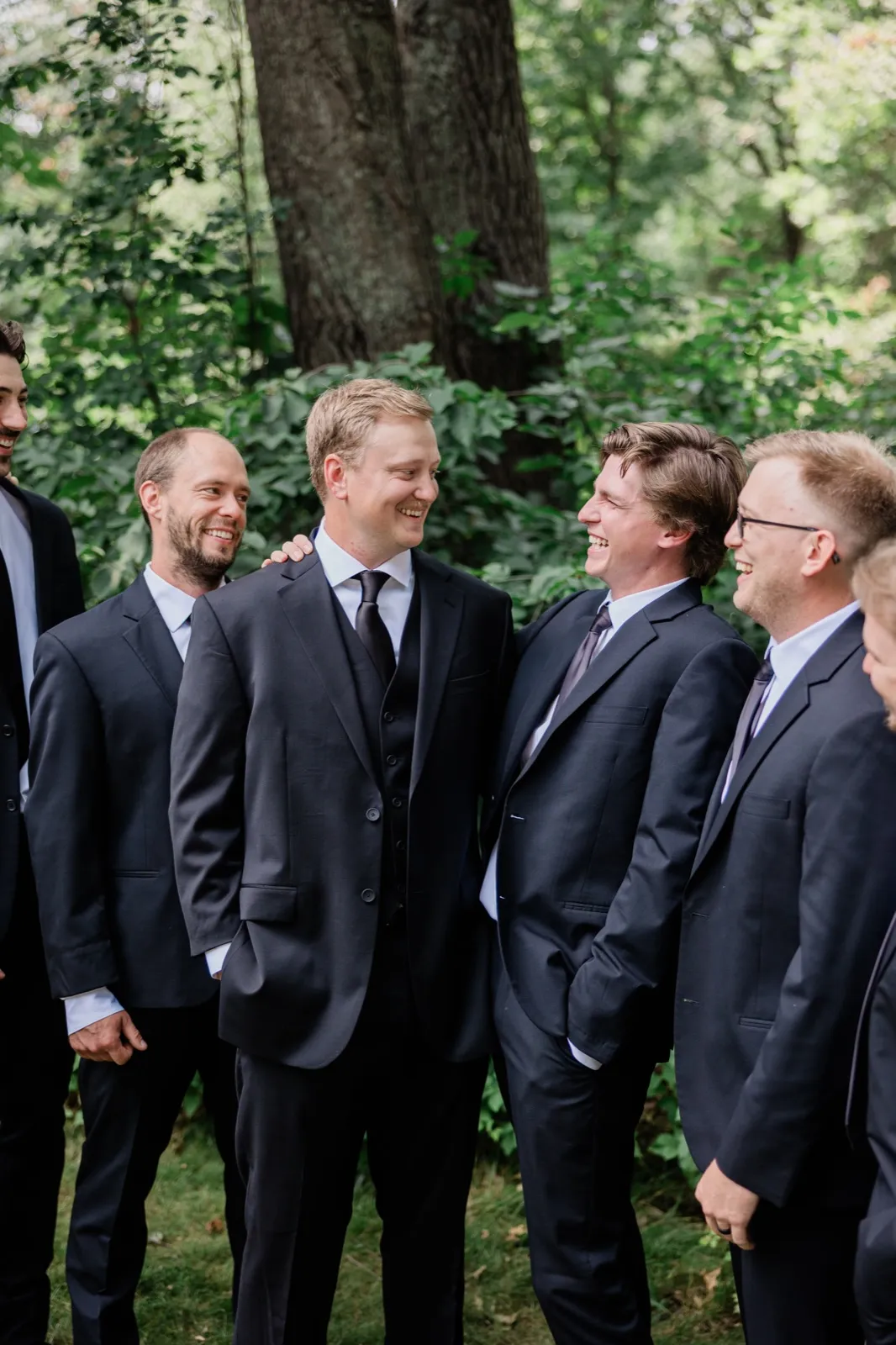 Carter laughs among five men in black suits who turn toward him, green summer woods behind them — Tim Larsen Photography, Brainerd Lakes MN