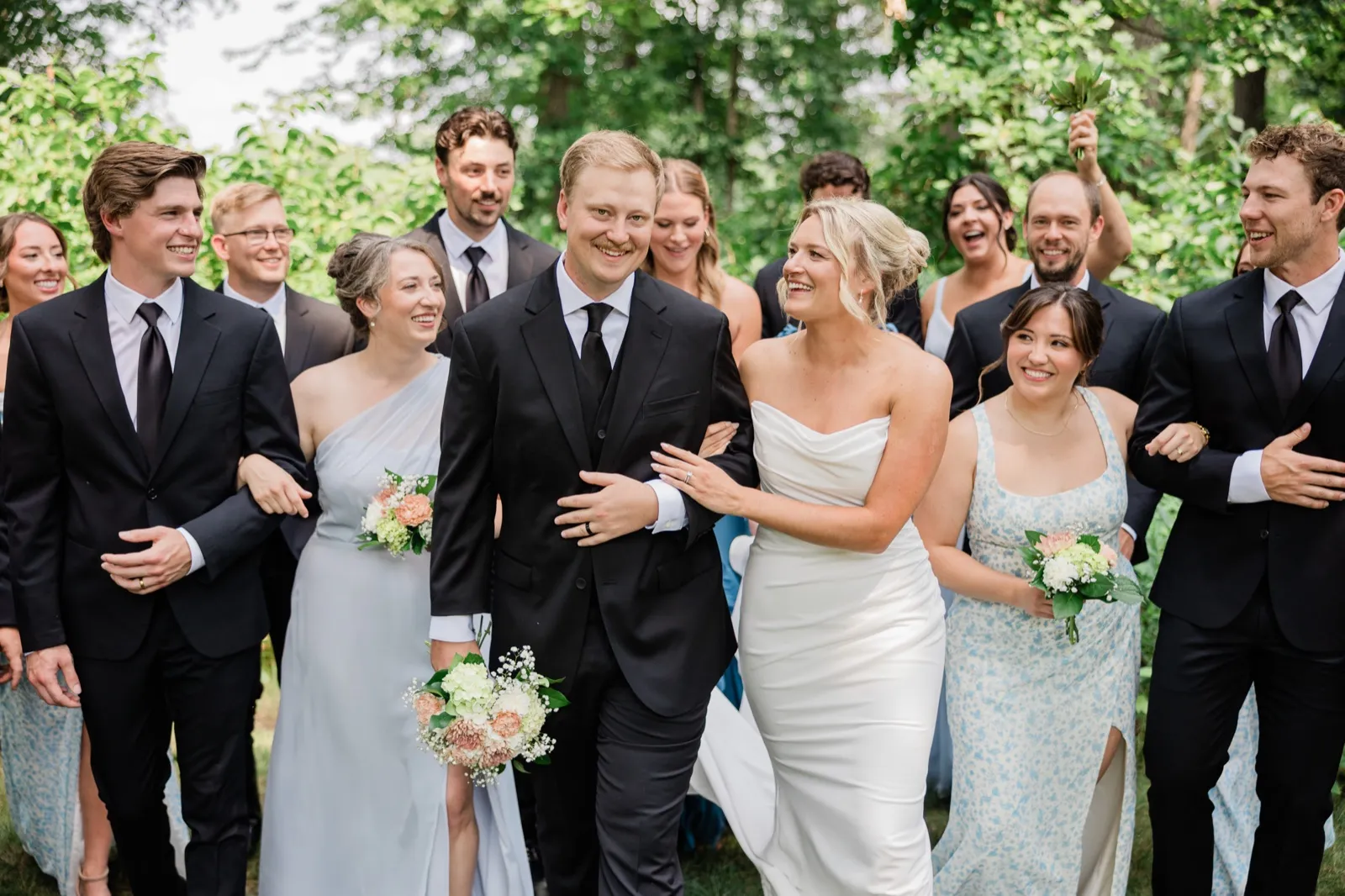 Amy and Carter walk arm in arm at the center of a large group of family and friends beneath a green canopy at Whitefish Lodge — Tim Larsen Photography, Brainerd Lakes MN