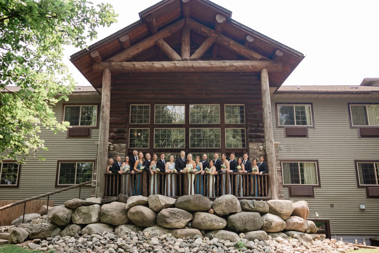 The whole group lines the log deck railing beneath the timber gable of Whitefish Lodge, a boulder retaining wall in the foreground — Tim Larsen Photography, Brainerd Lakes MN