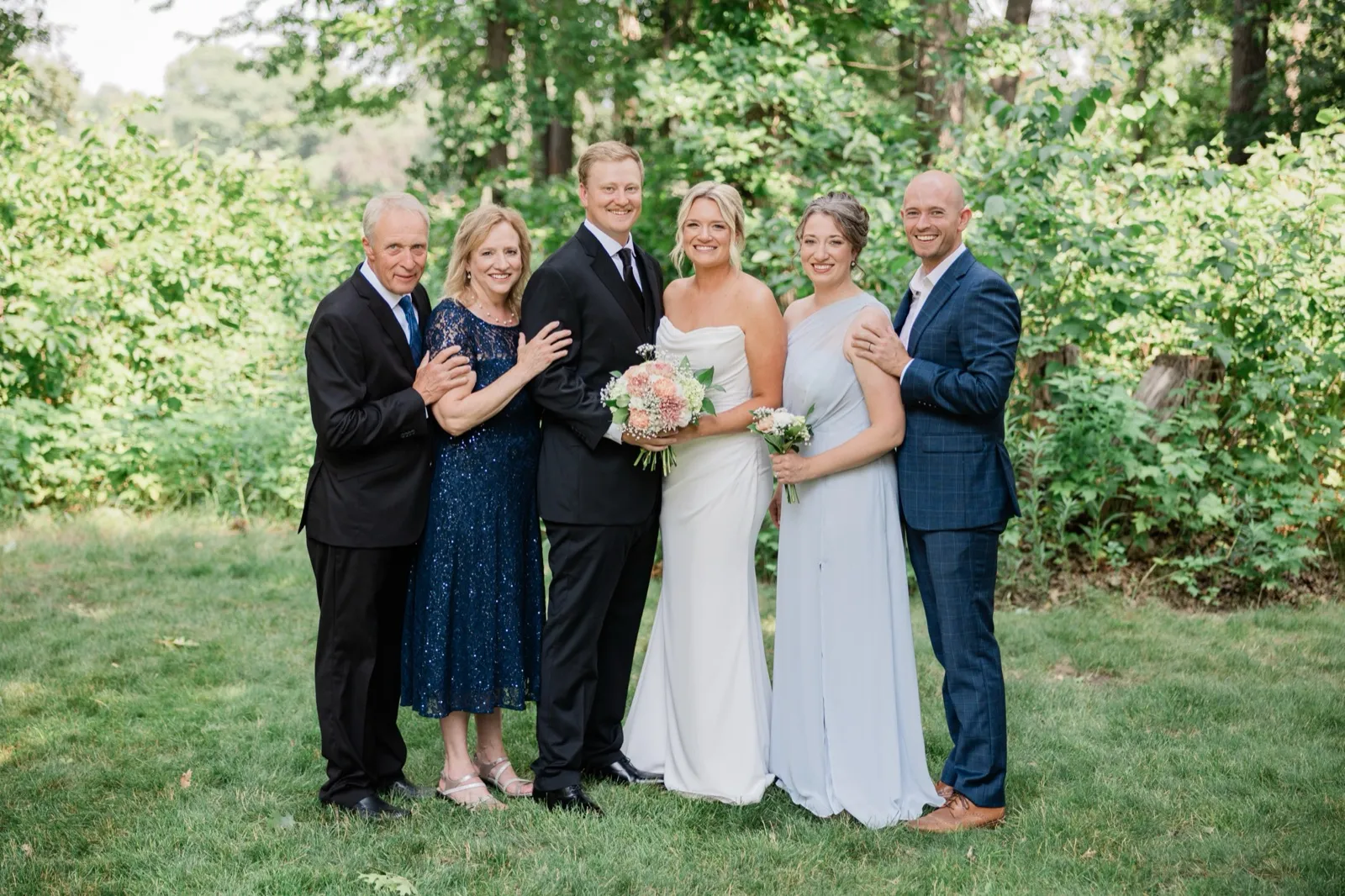Amy and Carter stand with family members on the grass lawn backed by dense summer greenery — Tim Larsen Photography, Brainerd Lakes MN