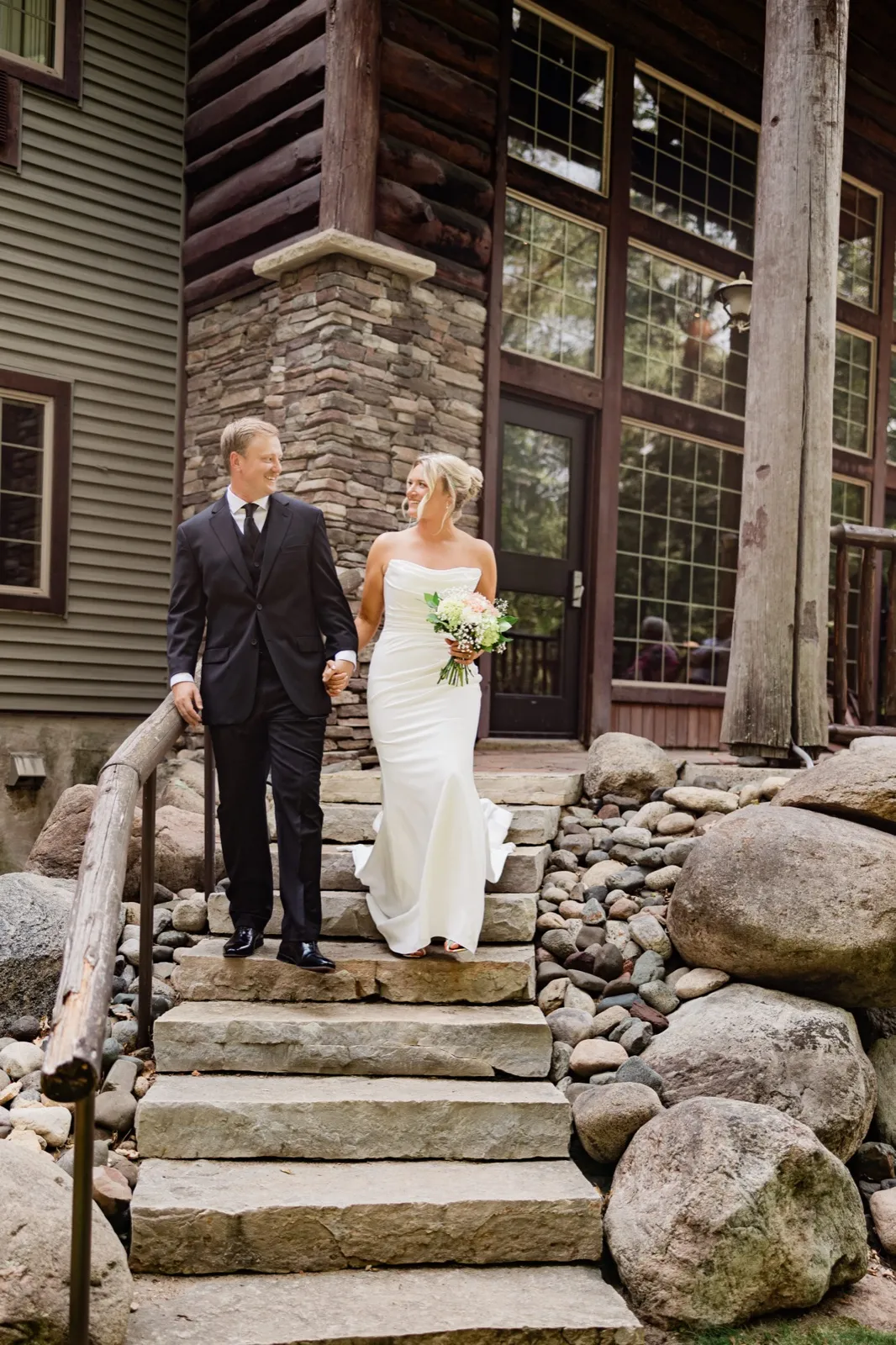 Amy and Carter hold hands walking down the wide stone steps outside the log-and-stone exterior of Whitefish Lodge — Tim Larsen Photography, Brainerd Lakes MN