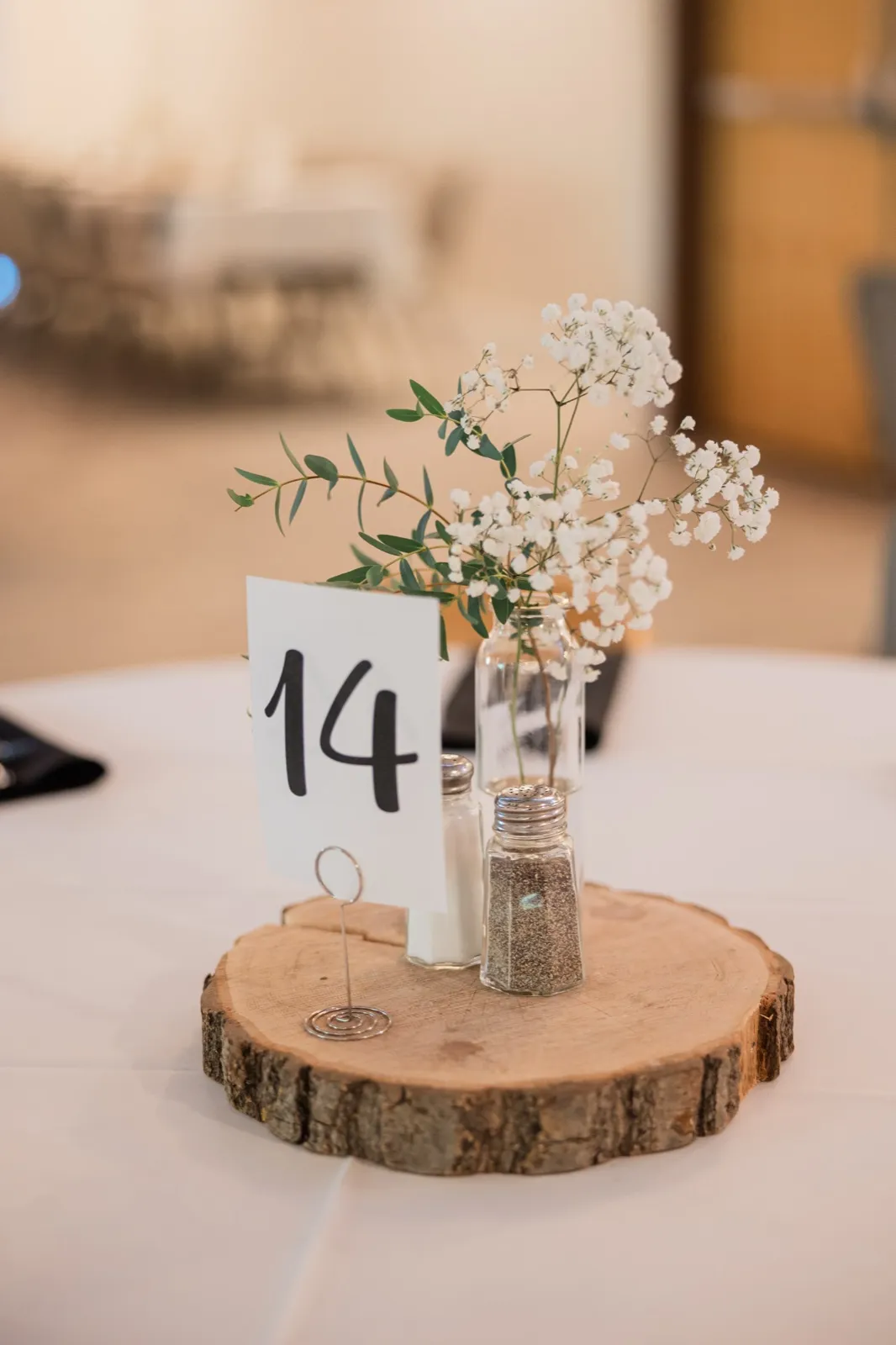 A reception centerpiece of baby's breath and eucalyptus in a glass bottle beside a table number on a wood slice — Tim Larsen Photography, Brainerd Lakes MN