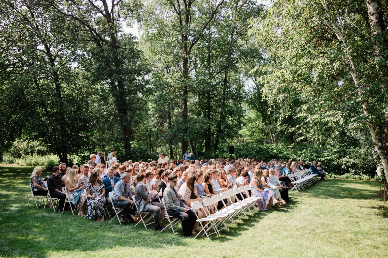 Guests fill rows of white folding chairs on the tree-shaded back lawn at Whitefish Lodge before the ceremony — Tim Larsen Photography, Brainerd Lakes MN