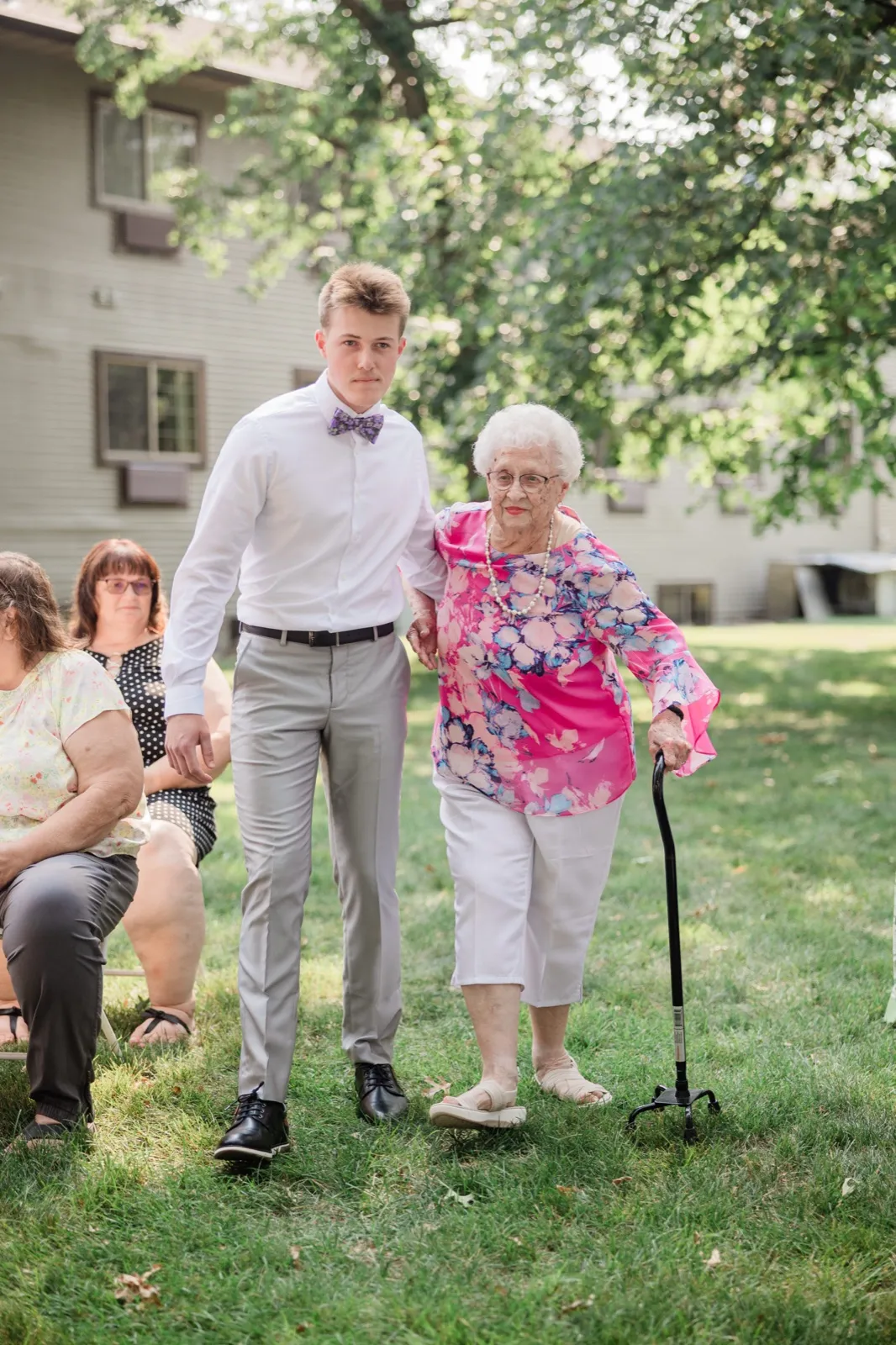 A young man in a bow tie steadies an older woman with a cane as they walk the grass aisle together — Tim Larsen Photography, Brainerd Lakes MN