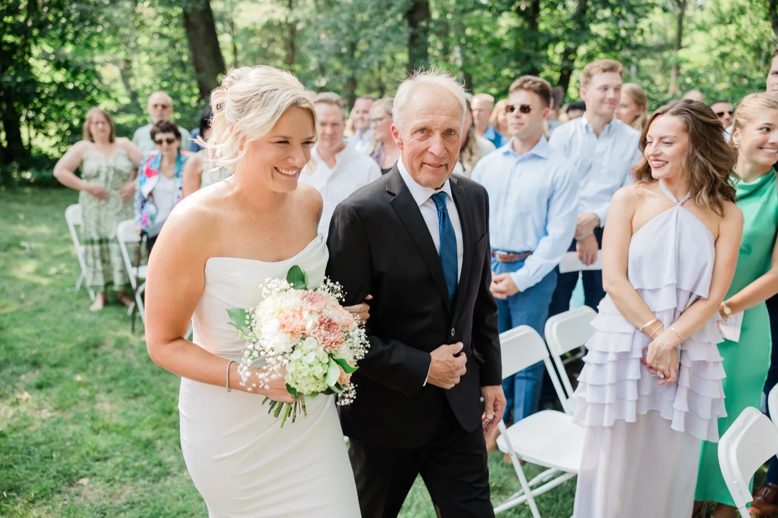 Amy smiles walking the grass aisle at Whitefish Lodge while standing guests in summer clothes turn to watch — Tim Larsen Photography, Brainerd Lakes MN