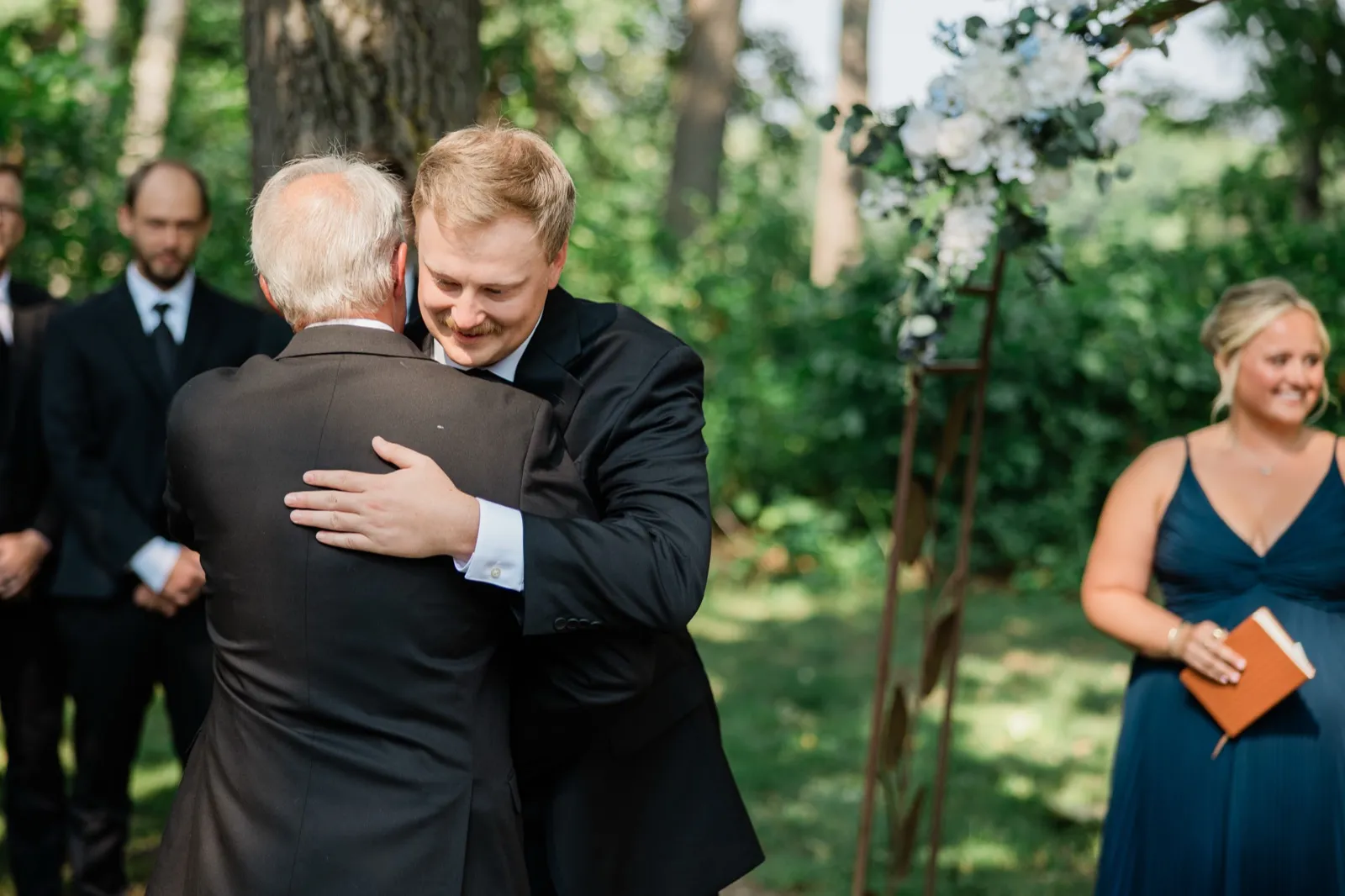 Carter embraces an older man in a dark suit at the front of the ceremony beside the floral arch — Tim Larsen Photography, Brainerd Lakes MN