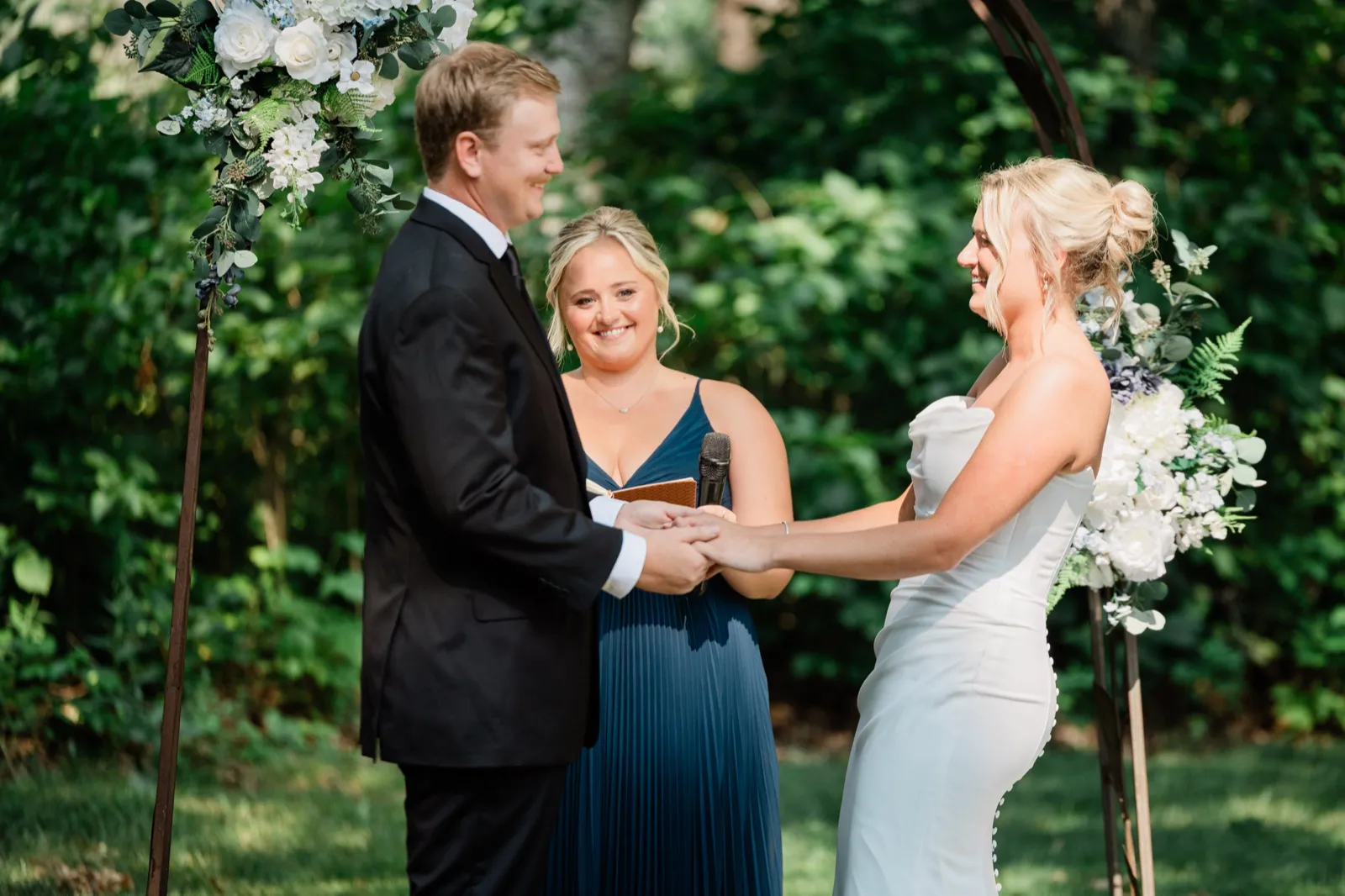 Amy and Carter hold hands beneath the metal arch while their friend, officiating in a navy dress, smiles between them — Tim Larsen Photography, Brainerd Lakes MN