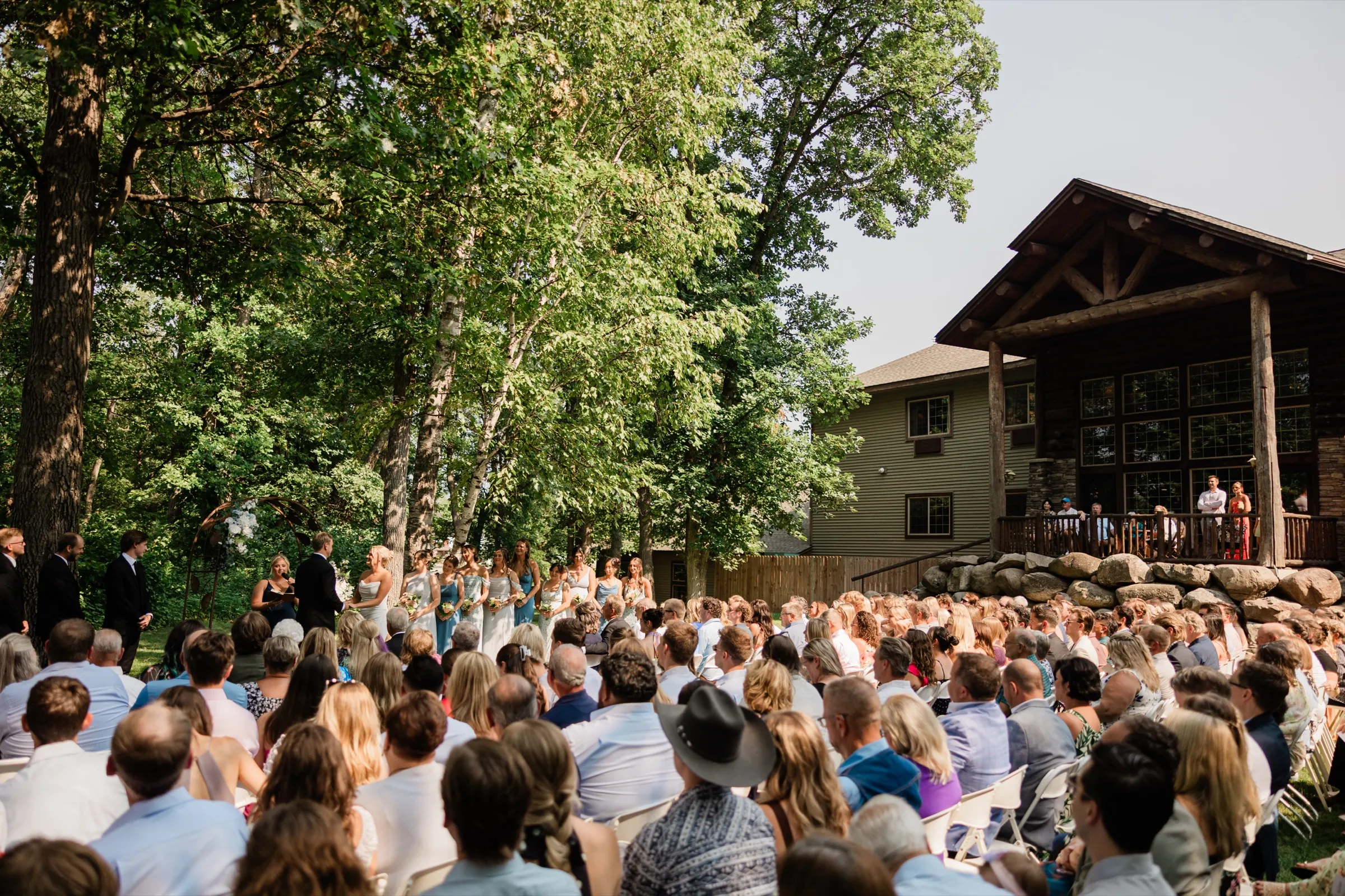 A large seated crowd watches Amy and Carter's ceremony on the tree-shaded lawn beside the timber-and-stone lodge — Tim Larsen Photography, Brainerd Lakes MN