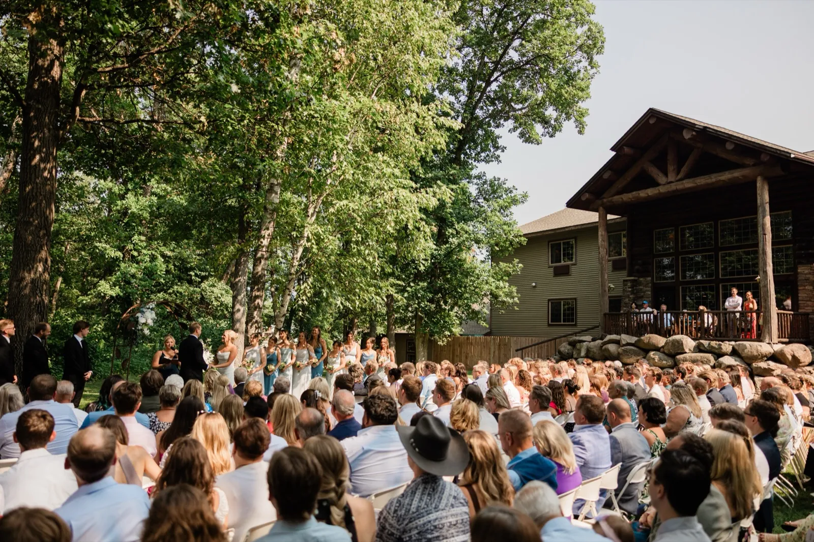 A large seated crowd watches Amy and Carter's ceremony on the tree-shaded lawn beside the timber-and-stone lodge — Tim Larsen Photography, Brainerd Lakes MN