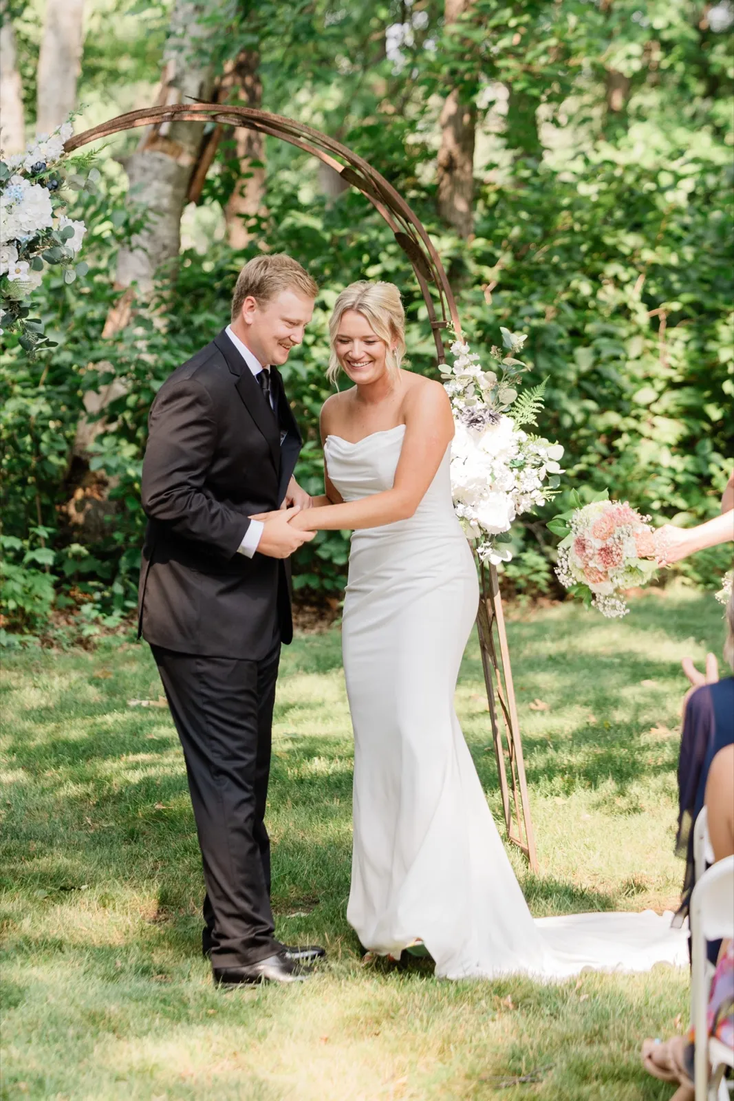 Amy laughs beside Carter under the rust-metal arch trimmed with white hydrangea, roses and ferns — Tim Larsen Photography, Brainerd Lakes MN
