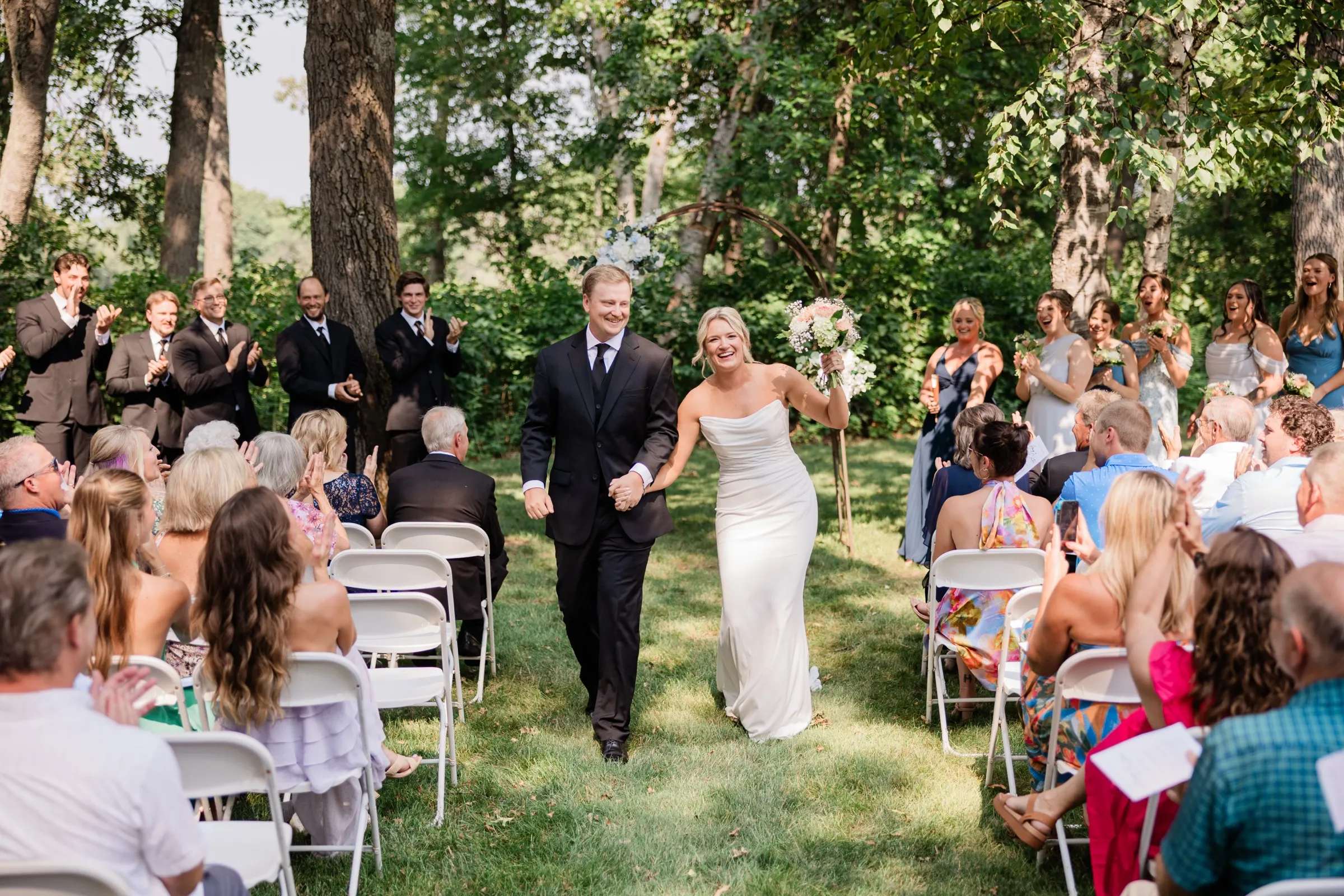 Amy raises her bouquet overhead as she and Carter walk back down the grass aisle married, guests applauding from white chairs — Tim Larsen Photography, Brainerd Lakes MN