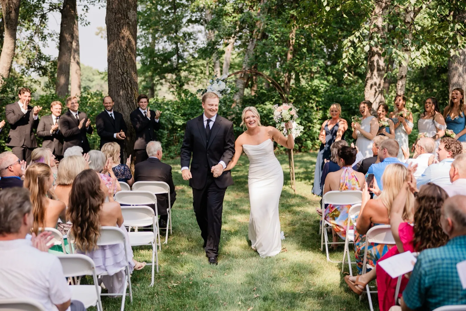 Amy raises her bouquet overhead as she and Carter walk back down the grass aisle married, guests applauding from white chairs — Tim Larsen Photography, Brainerd Lakes MN