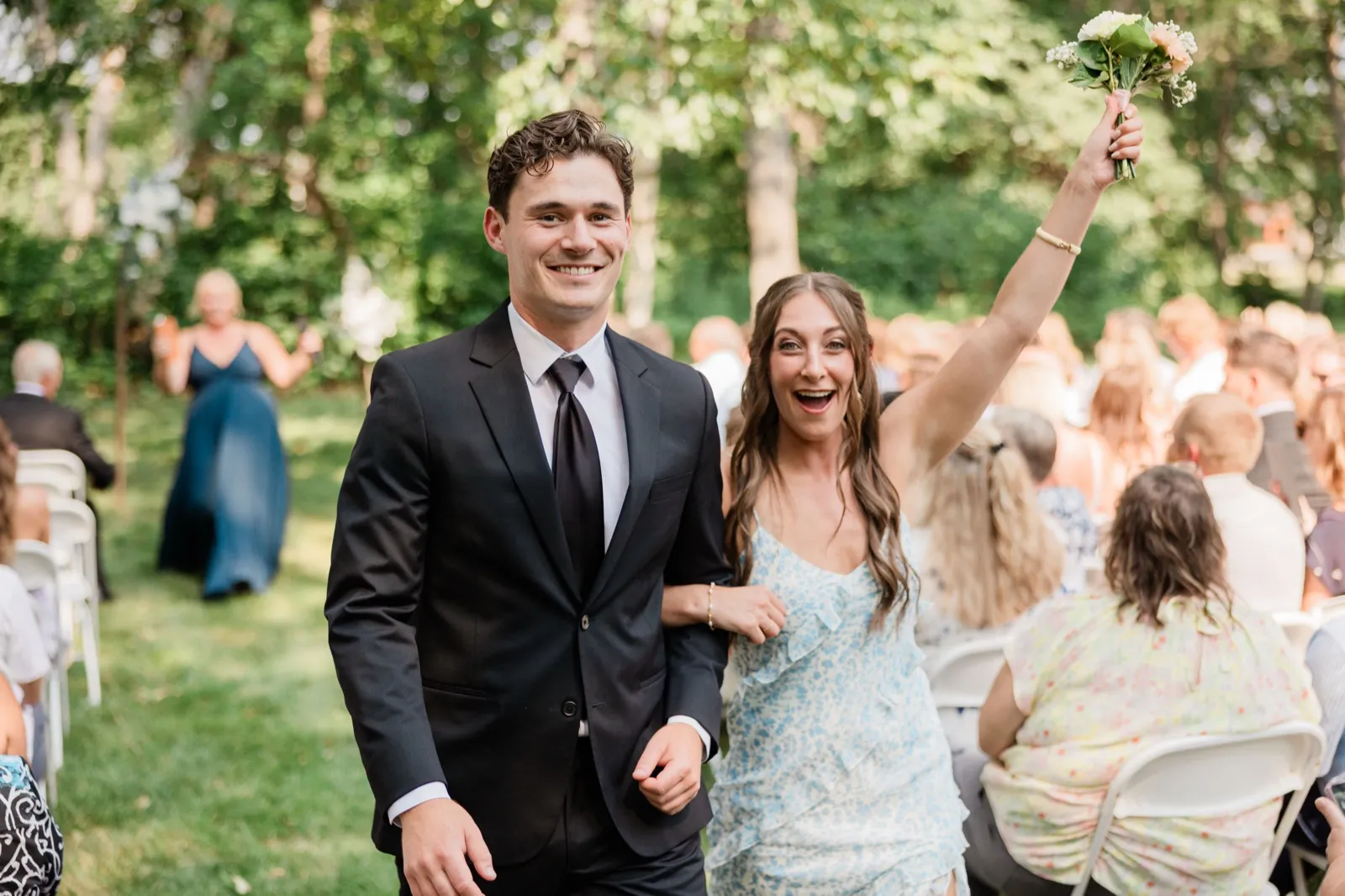 A guest in a blue floral dress throws an arm overhead walking back down the grass aisle arm in arm — Tim Larsen Photography, Brainerd Lakes MN