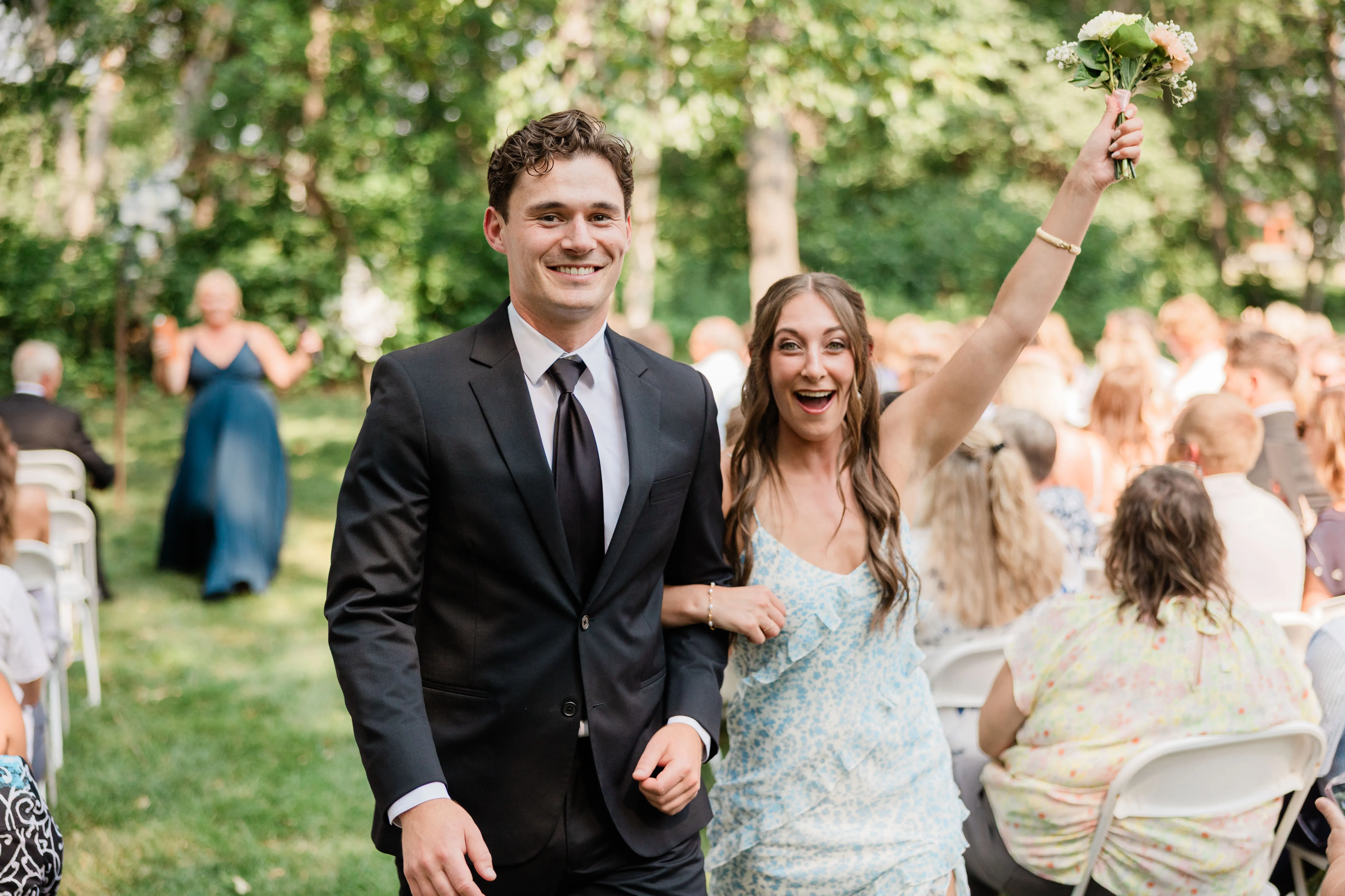 A guest in a blue floral dress throws an arm overhead walking back down the grass aisle arm in arm — Tim Larsen Photography, Brainerd Lakes MN
