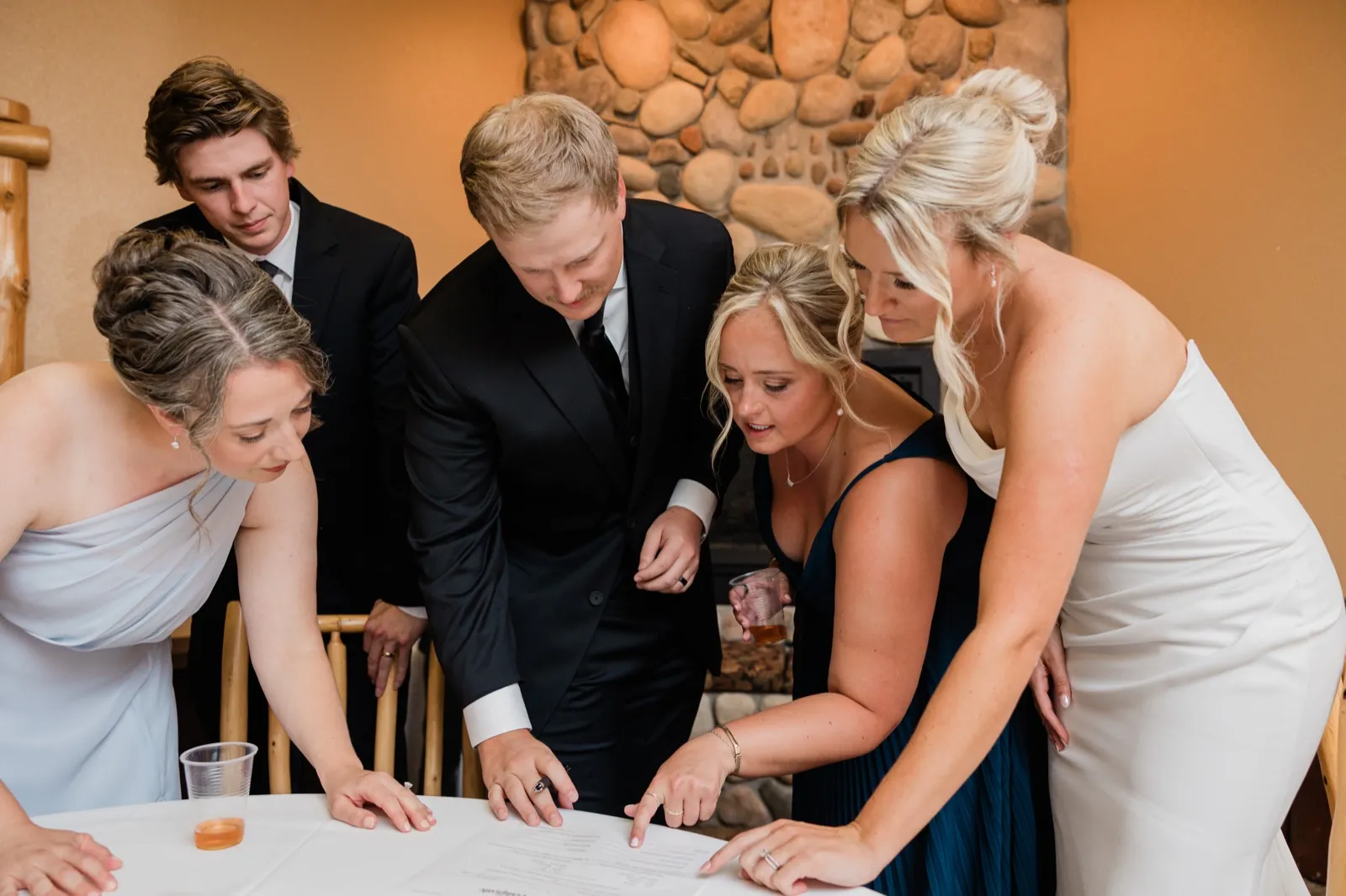 Carter signs the marriage license at a white-clothed table while Amy and three friends lean in and point, the river-rock fireplace behind them — Tim Larsen Photography, Brainerd Lakes MN