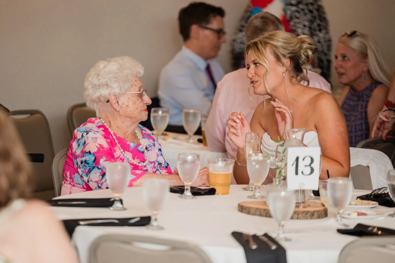 Amy leans in mid-conversation with an older woman in a pink floral dress at a round reception table — Tim Larsen Photography, Brainerd Lakes MN