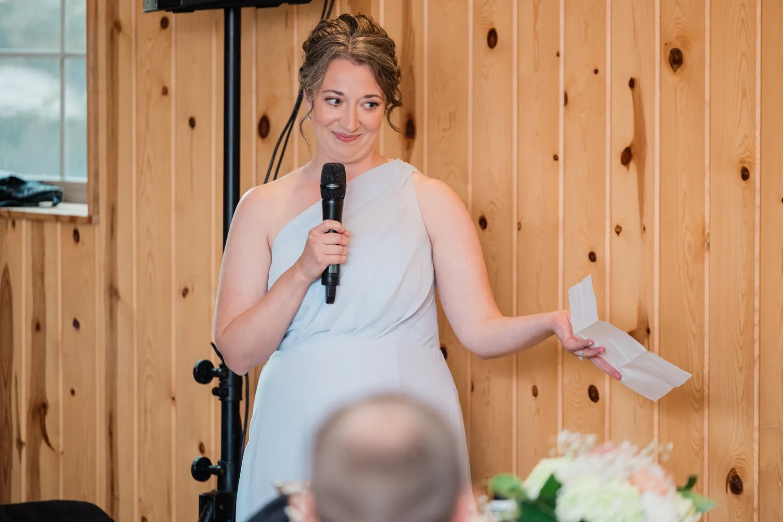 A friend in a pale blue one-shoulder dress holds a microphone and her notes during a toast in the pine-paneled room — Tim Larsen Photography, Brainerd Lakes MN