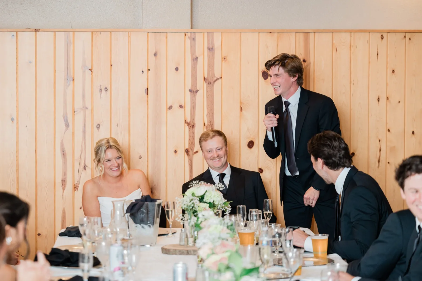 A young man laughs mid-toast while Amy and Carter laugh along at the head table against the pine-paneled wall — Tim Larsen Photography, Brainerd Lakes MN