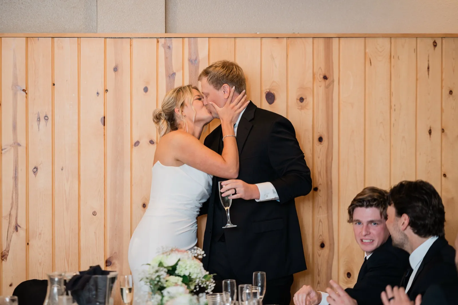 Amy pulls Carter in for a kiss against the knotty pine wall at the head table while a friend mugs at the camera — Tim Larsen Photography, Brainerd Lakes MN