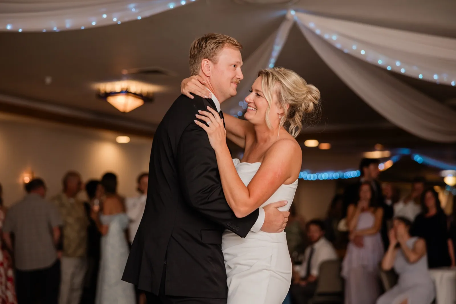 Amy smiles up at Carter with her arms around his neck during their first dance beneath draped fabric and blue string lights — Tim Larsen Photography, Brainerd Lakes MN