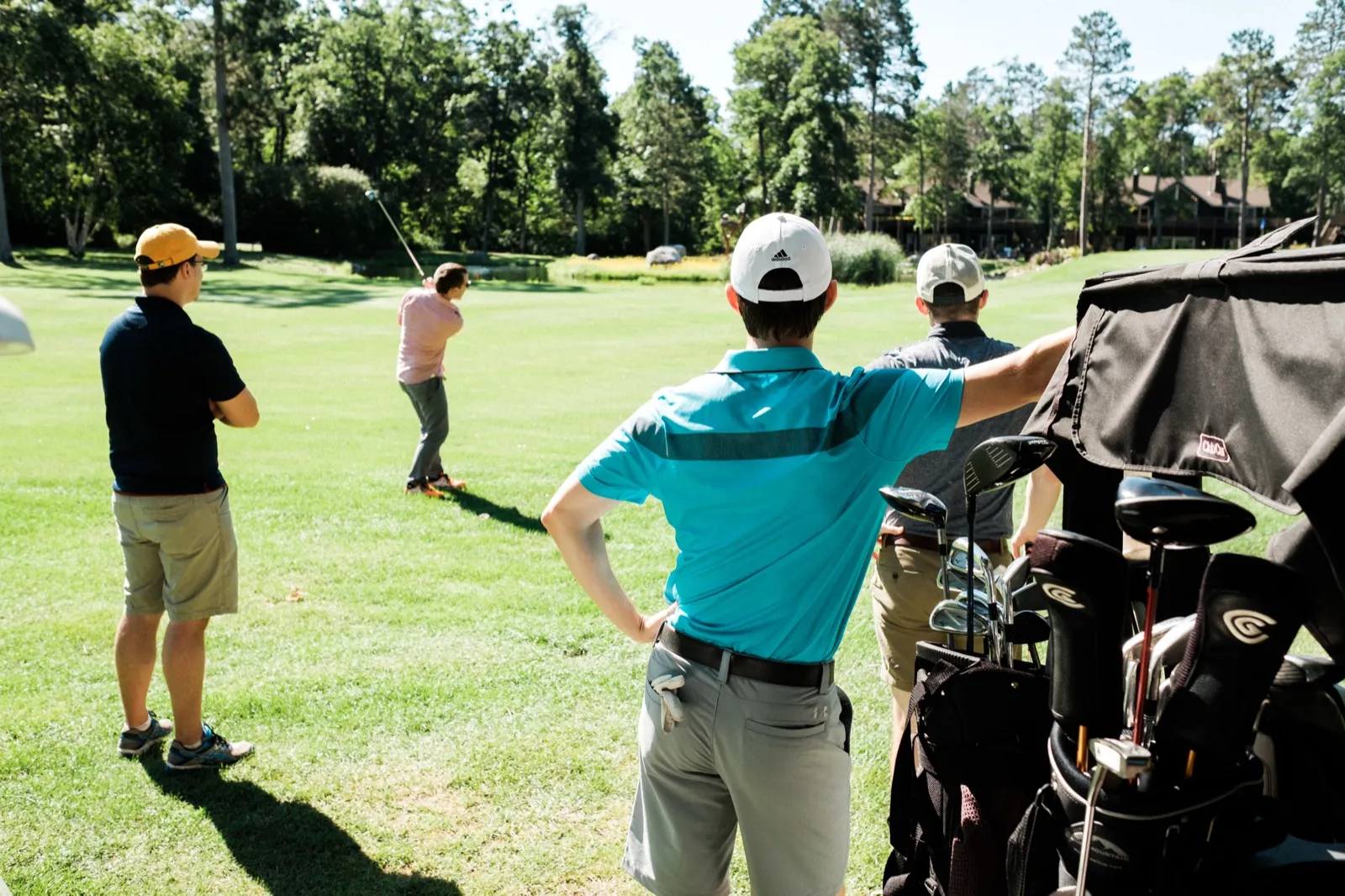 Three men in golf attire watch a fourth swing a club on a sunlit fairway, with a golf bag full of clubs framing the foreground and the Grand View Lodge buildings visible through the trees. — Tim Larsen Photography, Brainerd Lakes MN