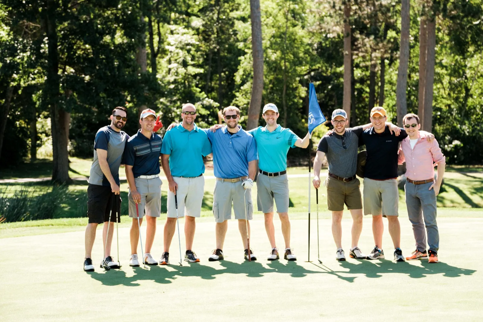 A group of eight men in polos and shorts pose together on a sunlit golf green, one holding a Grand View Lodge flag. — Tim Larsen Photography, Brainerd Lakes MN