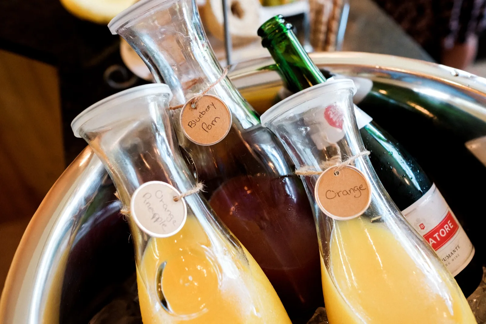 A silver tray holds glass carafes of orange and blueberry-pomegranate juice with handwritten kraft-paper tags beside a green bottle of sparkling wine. — Tim Larsen Photography, Brainerd Lakes MN