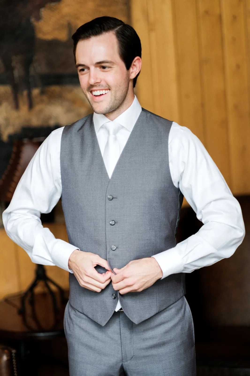 The groom laughs while buttoning his gray vest in a wood-paneled room with a dark landscape painting behind him. — Tim Larsen Photography, Brainerd Lakes MN