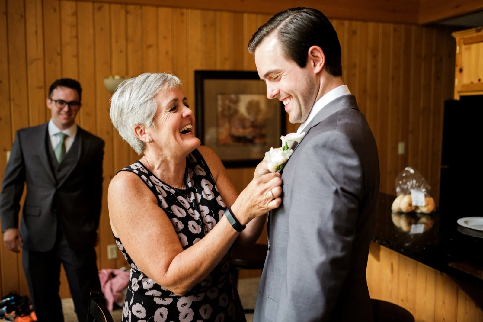 A woman laughs as she pins a white boutonniere to the groom's gray suit lapel while a groomsman watches from the background in a wood-paneled room. — Tim Larsen Photography, Brainerd Lakes MN