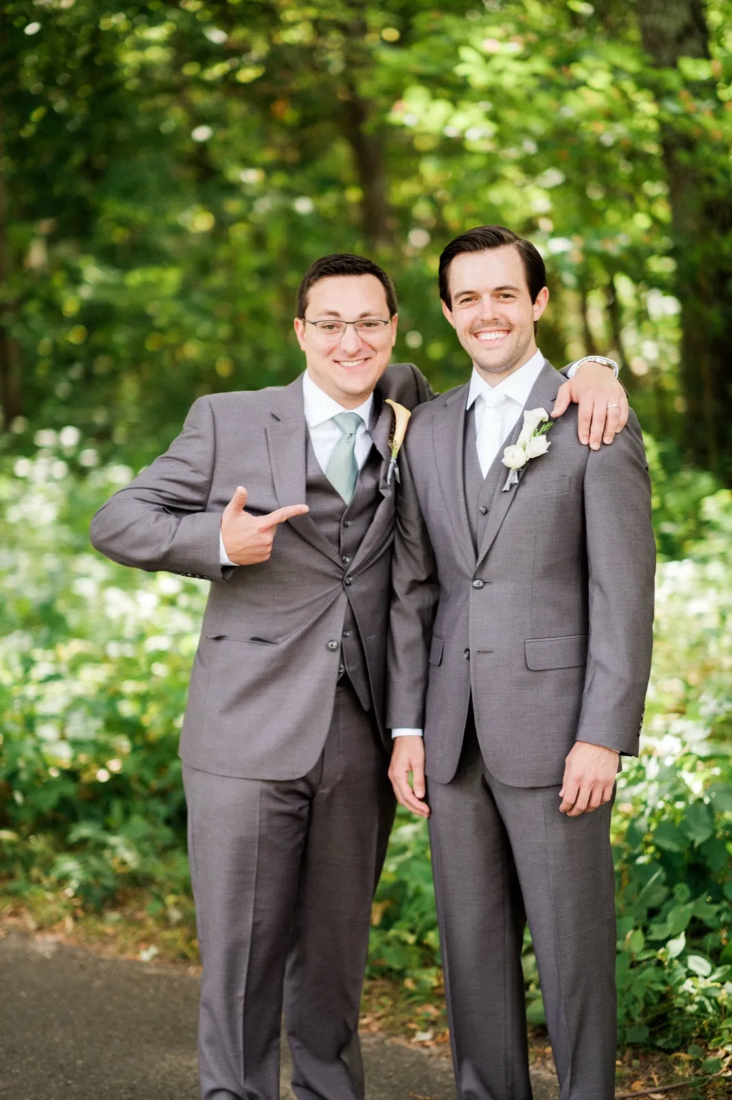 The groom and his groomsman stand arm-in-arm on a wooded path in matching gray suits, smiling at the camera as the groomsman playfully points toward the groom. — Tim Larsen Photography, Brainerd Lakes MN