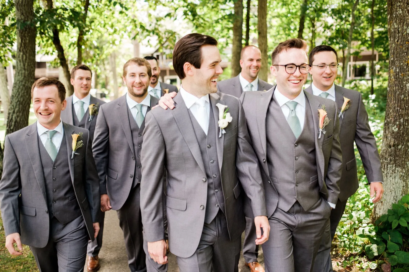 The groom laughs with his groomsmen as they walk together along a wooded path in matching gray suits with sage green ties. — Tim Larsen Photography, Brainerd Lakes MN