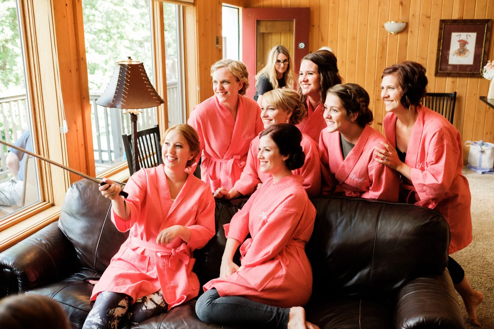 The bride and her six bridesmaids in matching coral robes crowd onto a leather sofa as the bride extends a selfie stick to photograph the group. — Tim Larsen Photography, Brainerd Lakes MN