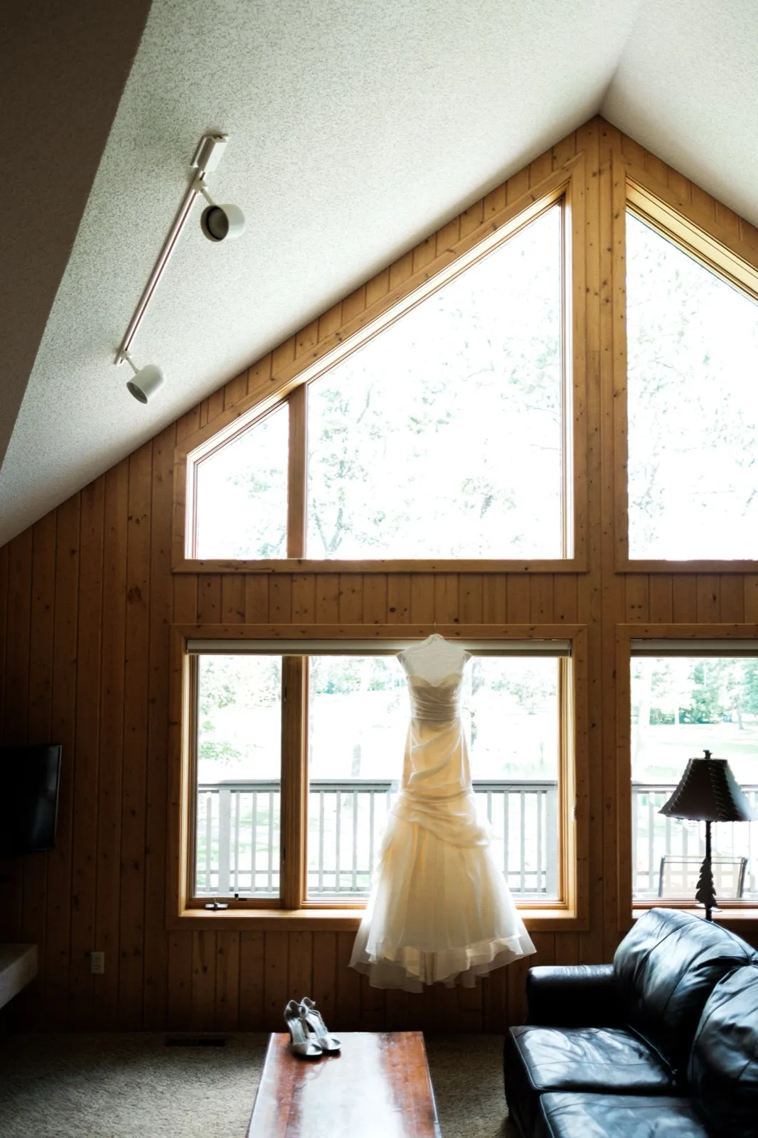 A wedding dress hangs in the peak of a tall A-frame window inside a wood-paneled cabin, with bridal shoes resting on the coffee table below. — Tim Larsen Photography, Brainerd Lakes MN