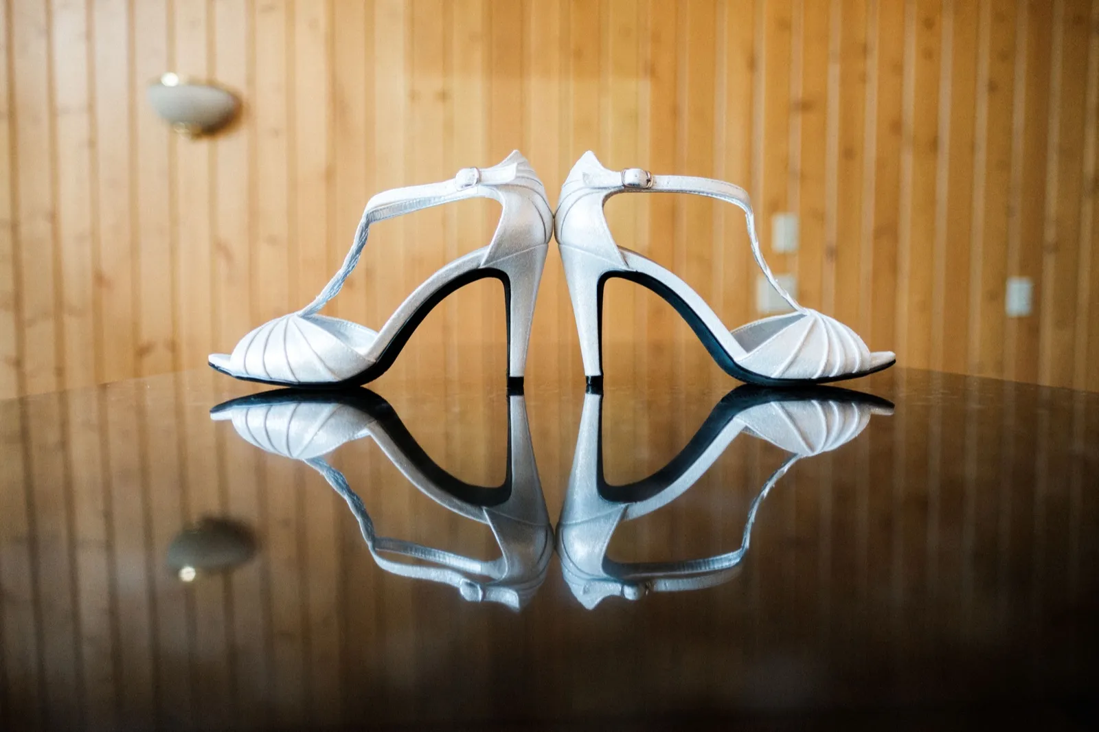 A pair of white peep-toe bridal heels stand on a glossy table, mirrored in its reflection, against a warm wood-paneled wall. — Tim Larsen Photography, Brainerd Lakes MN