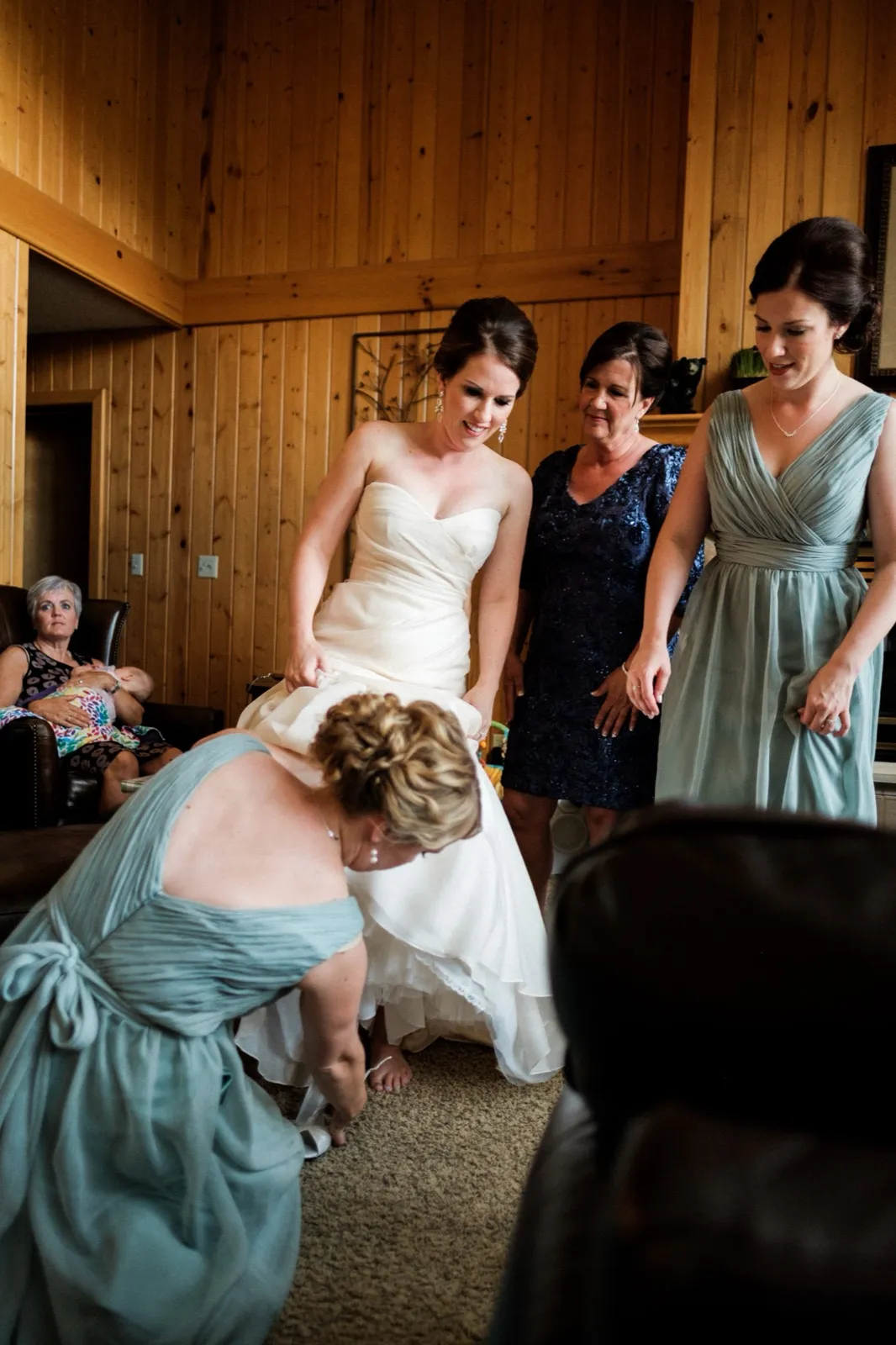 A bridesmaid kneels to adjust the hem of the bride's gown while the bride's mother and another bridesmaid look on, with a grandmother holding a baby visible in the background. — Tim Larsen Photography, Brainerd Lakes MN