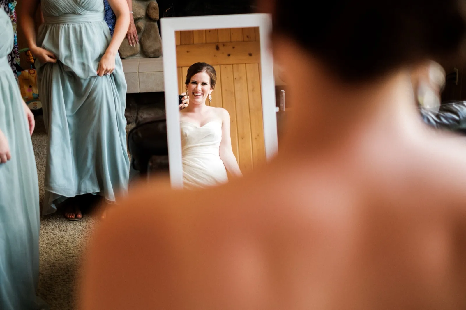 The bride laughs in a standing mirror's reflection, framed between an out-of-focus bridesmaid and another figure in the foreground. — Tim Larsen Photography, Brainerd Lakes MN