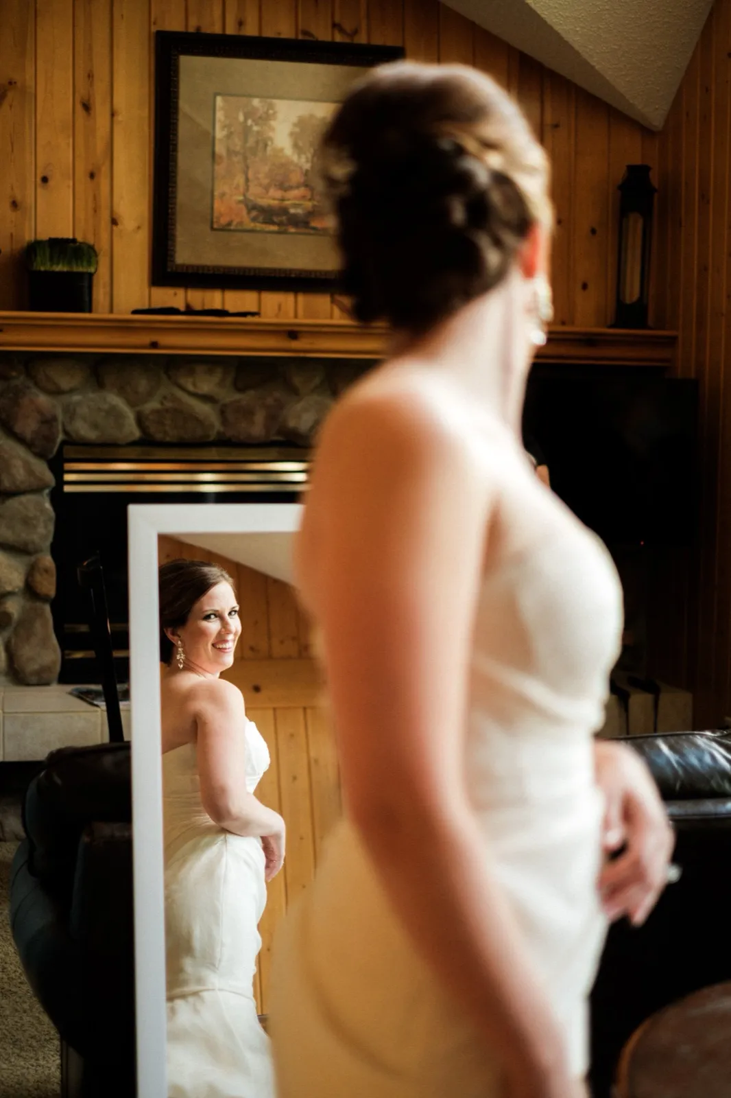 The bride stands out of focus in the foreground while her smiling reflection appears in a framed mirror behind her in a wood-paneled cabin room. — Tim Larsen Photography, Brainerd Lakes MN